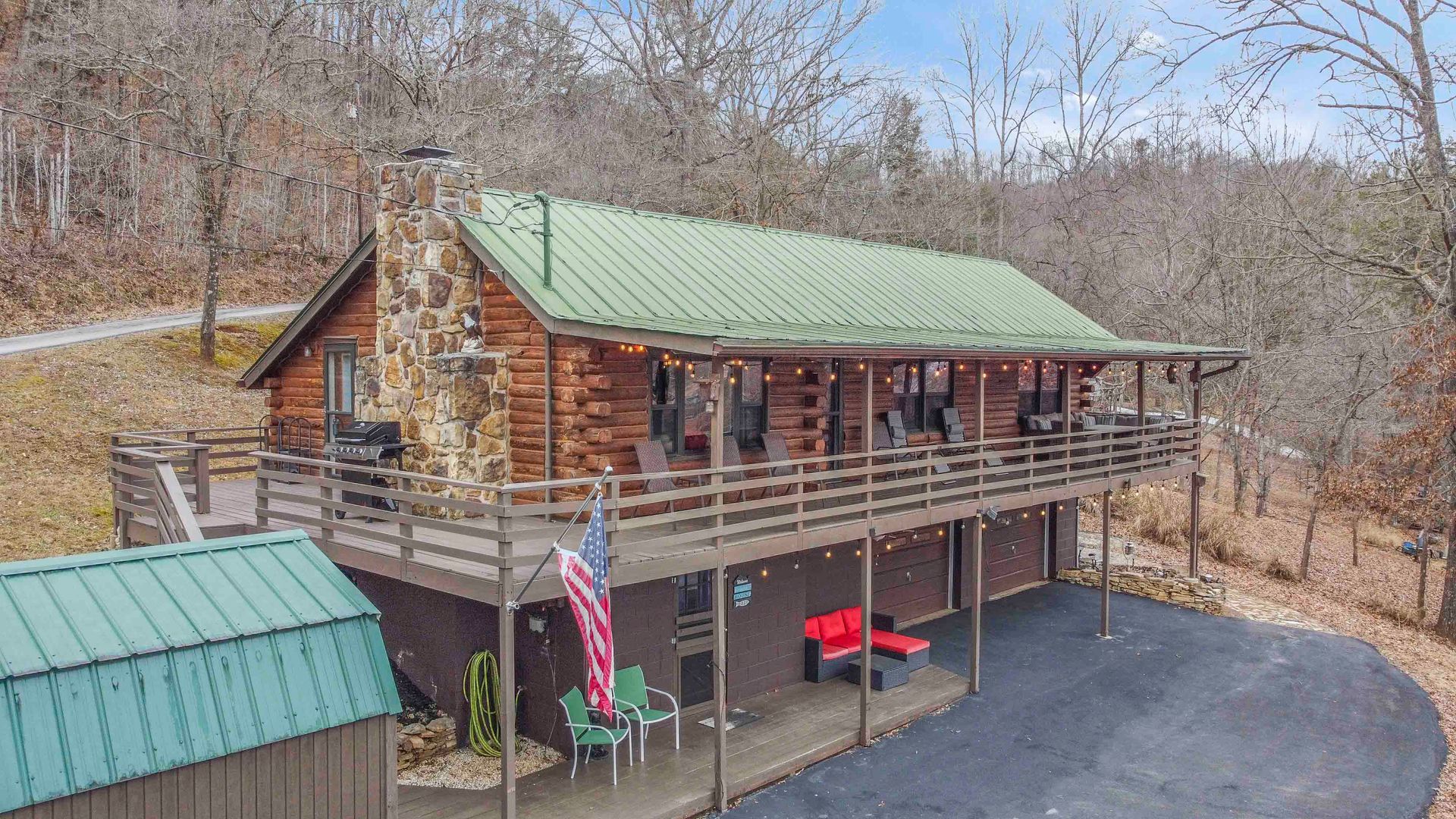 Log cabin with a wrap-around porch and green roof, in a wooded setting.
