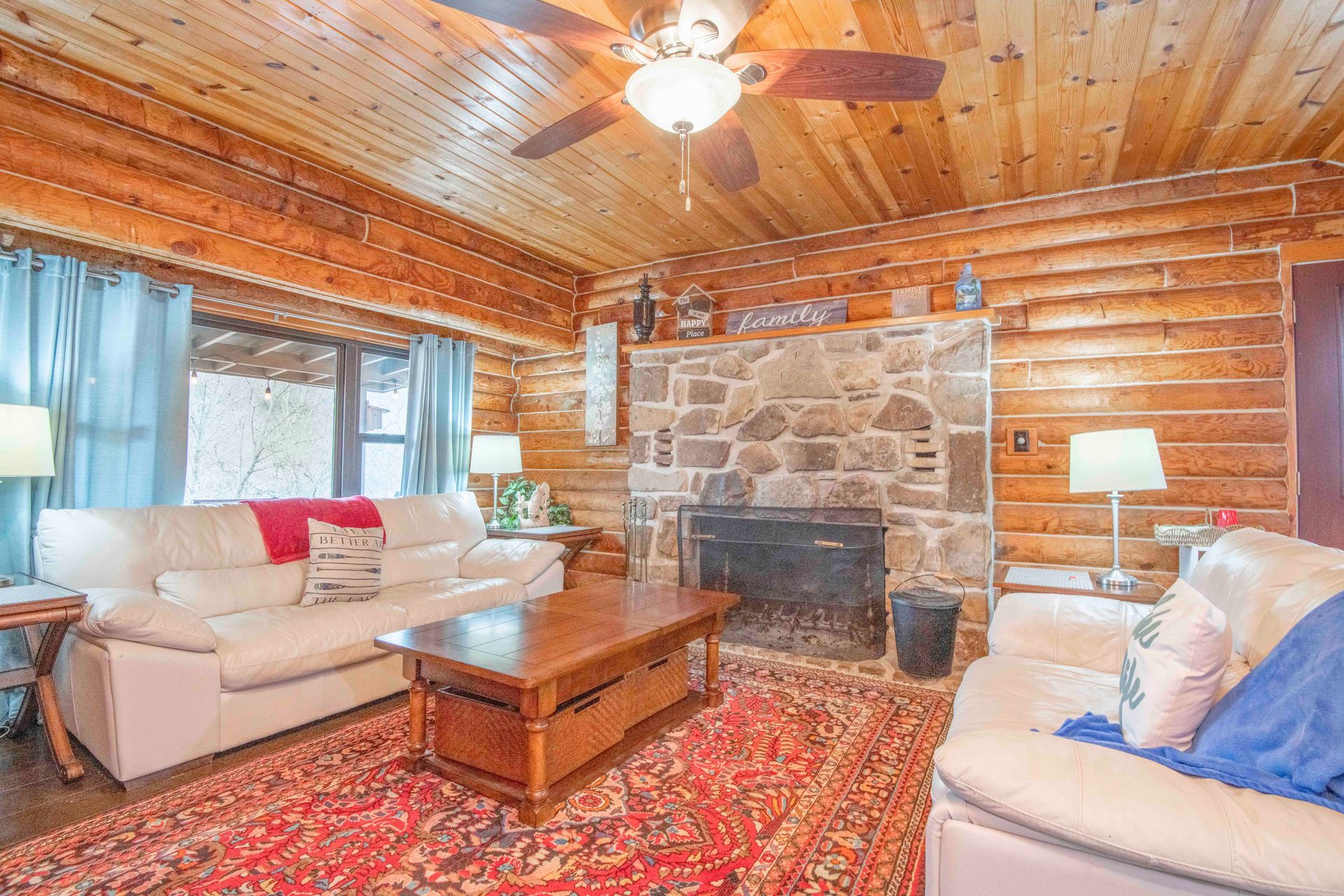 Cozy living room with a stone fireplace, wood paneling, two white sofas, and a red rug.