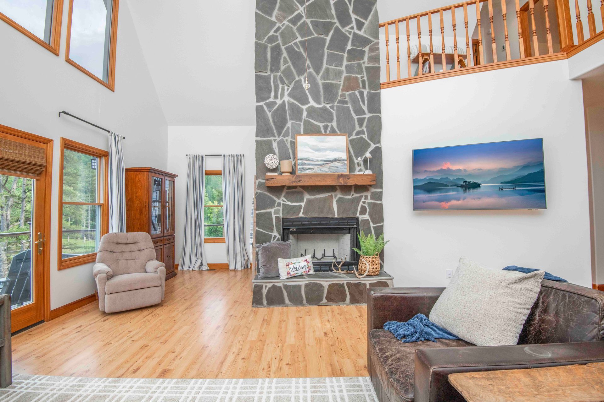 Living room with stone fireplace, wooden floors, and large windows overlooking the outdoors.