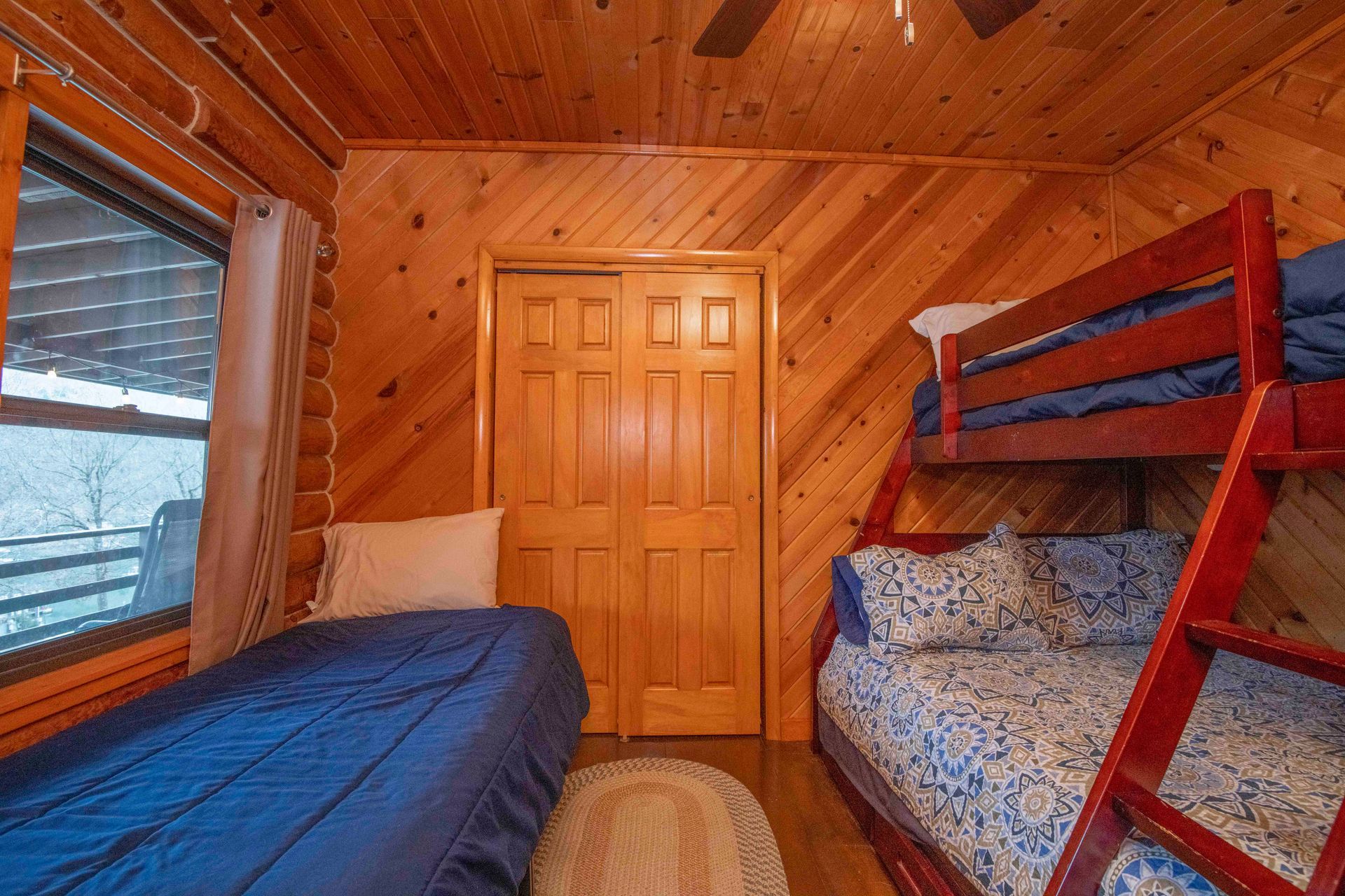 A small bedroom with a bunk bed and a single bed, both covered in blue bedding, wooden walls.