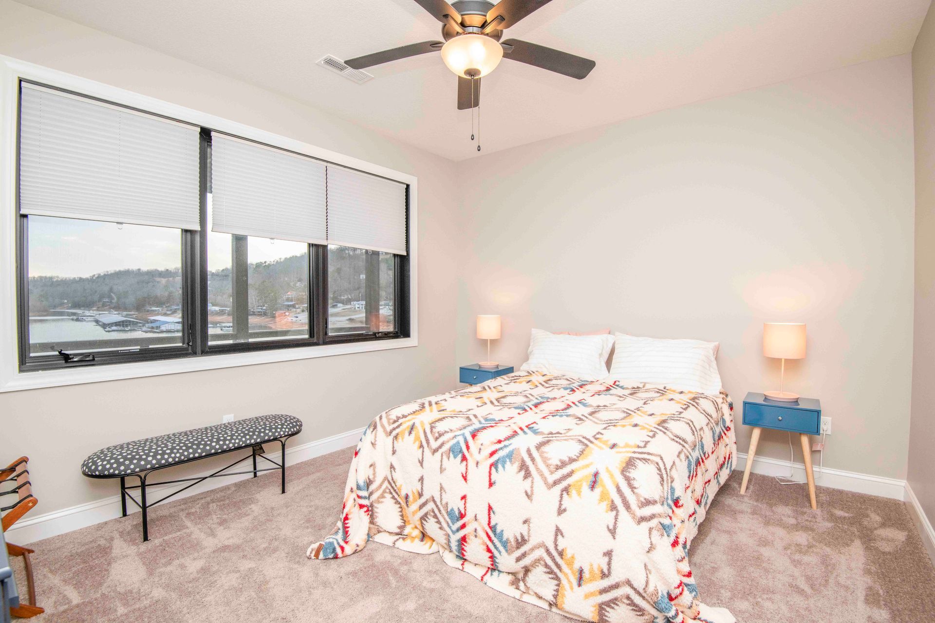 Bedroom with a bed, bench, blue nightstands, and windows with a view of a city.
