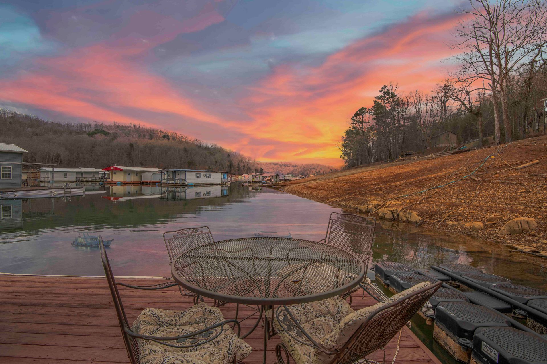 Sunset over a lake, a metal table and chairs on a dock. Houses on the far side, trees on the shores.