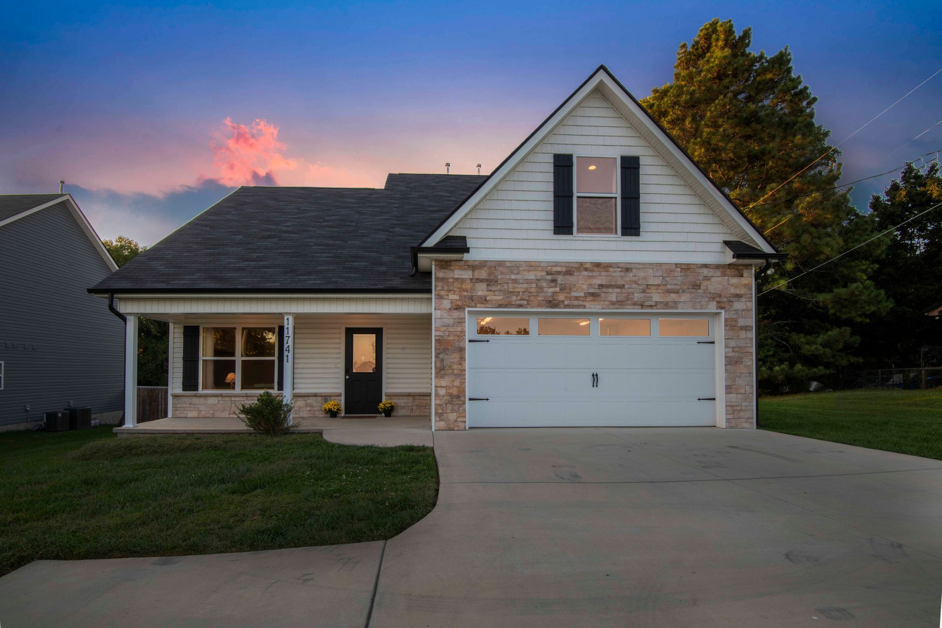 House with white siding, stone accents, black shutters, and a two-car garage. Overcast sky at dusk.