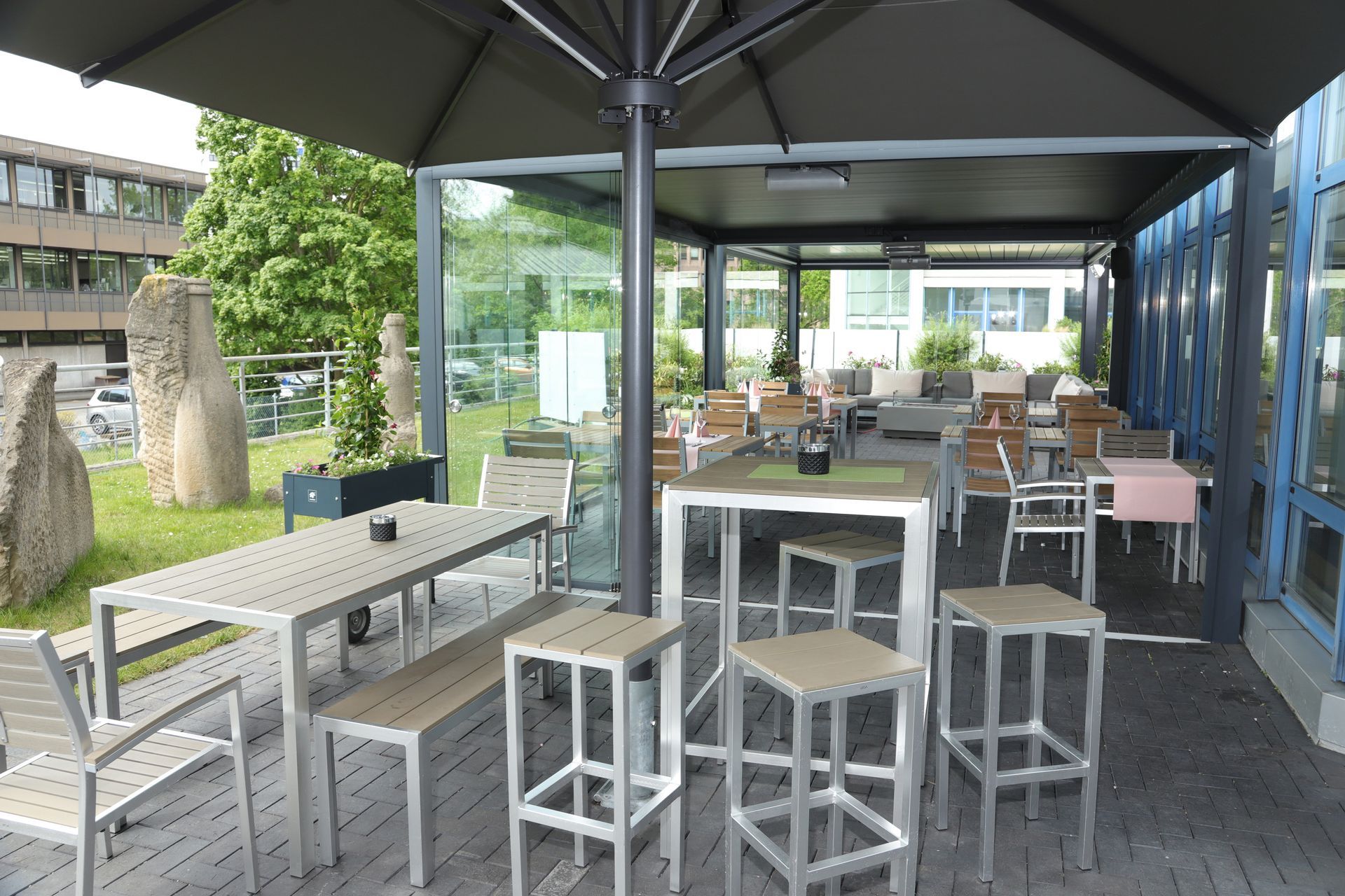 Sitzgelegenheiten im Freien unter einem großen Sonnenschirm mit Tischen, Hockern und Glaswänden neben einem Gartenweg.