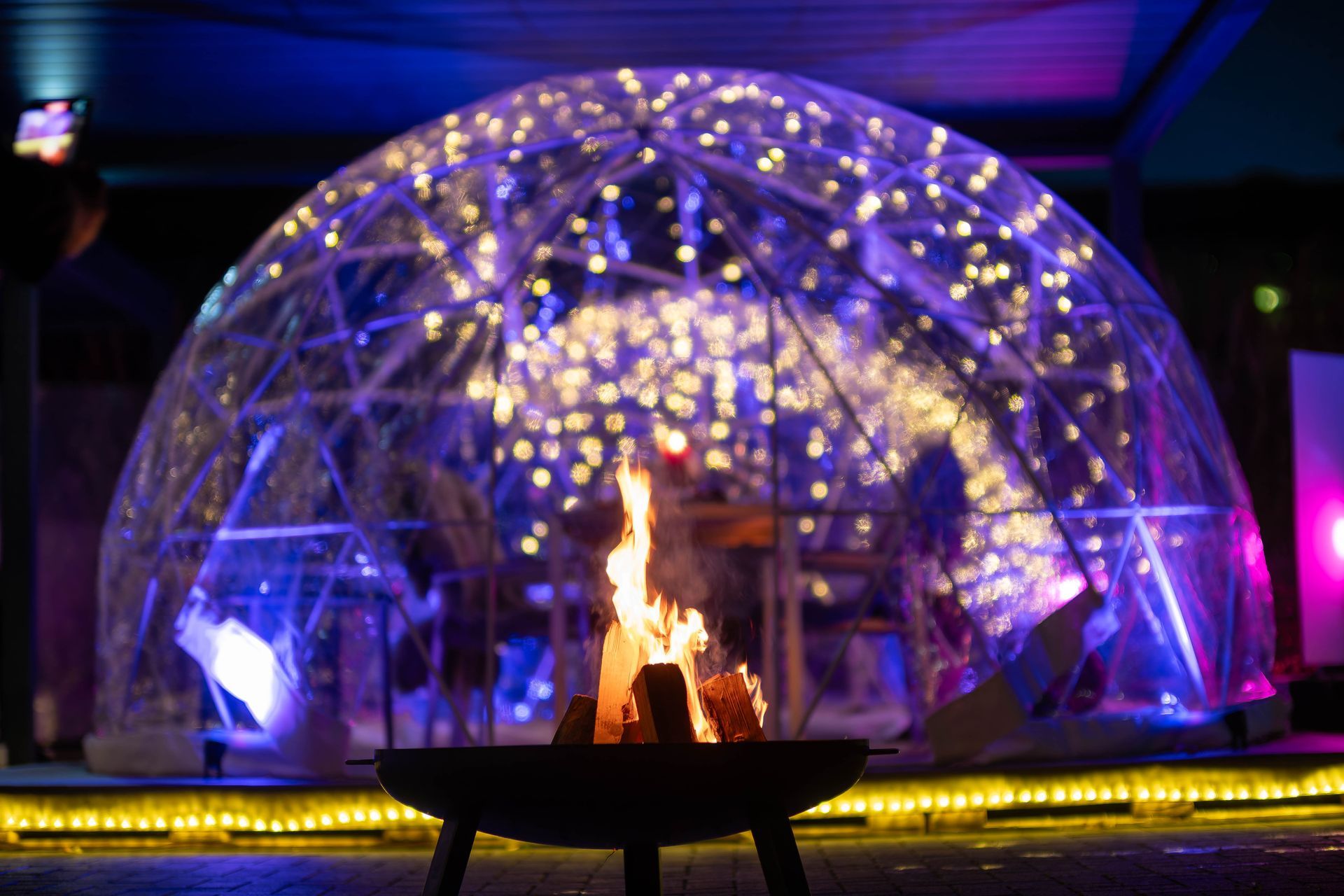 Feuerstelle im Inneren einer leuchtenden, transparenten Kuppel bei Nacht, blau und violett beleuchtet.