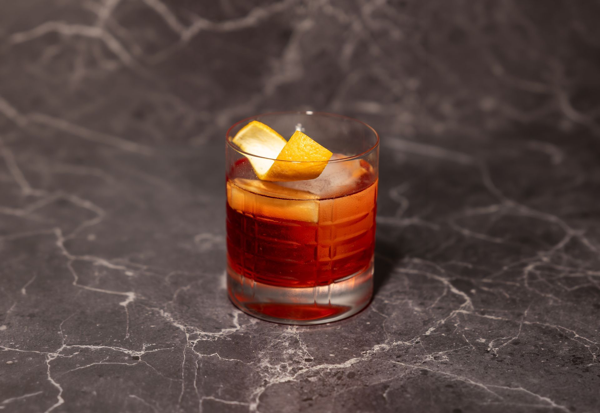 Kurzer, bernsteinfarbener Cocktail in einem niedrigen Glas mit Orangenschalengarnitur auf einer dunklen Marmoroberfläche