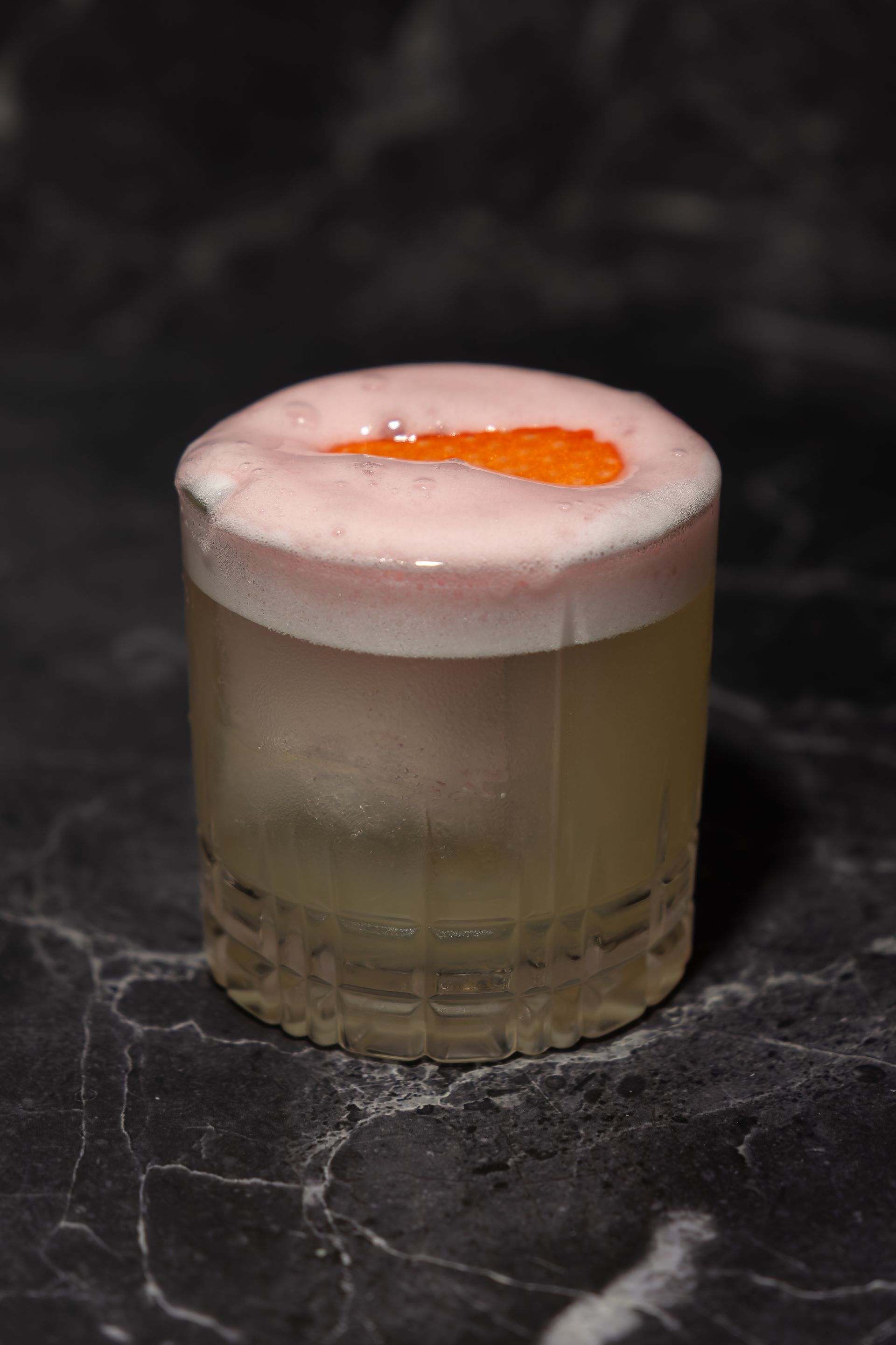 Ein schaumiger Cocktail in einem durchsichtigen Lowball-Glas mit einer Orangenscheibe als Garnitur auf einer dunklen Marmoroberfläche.