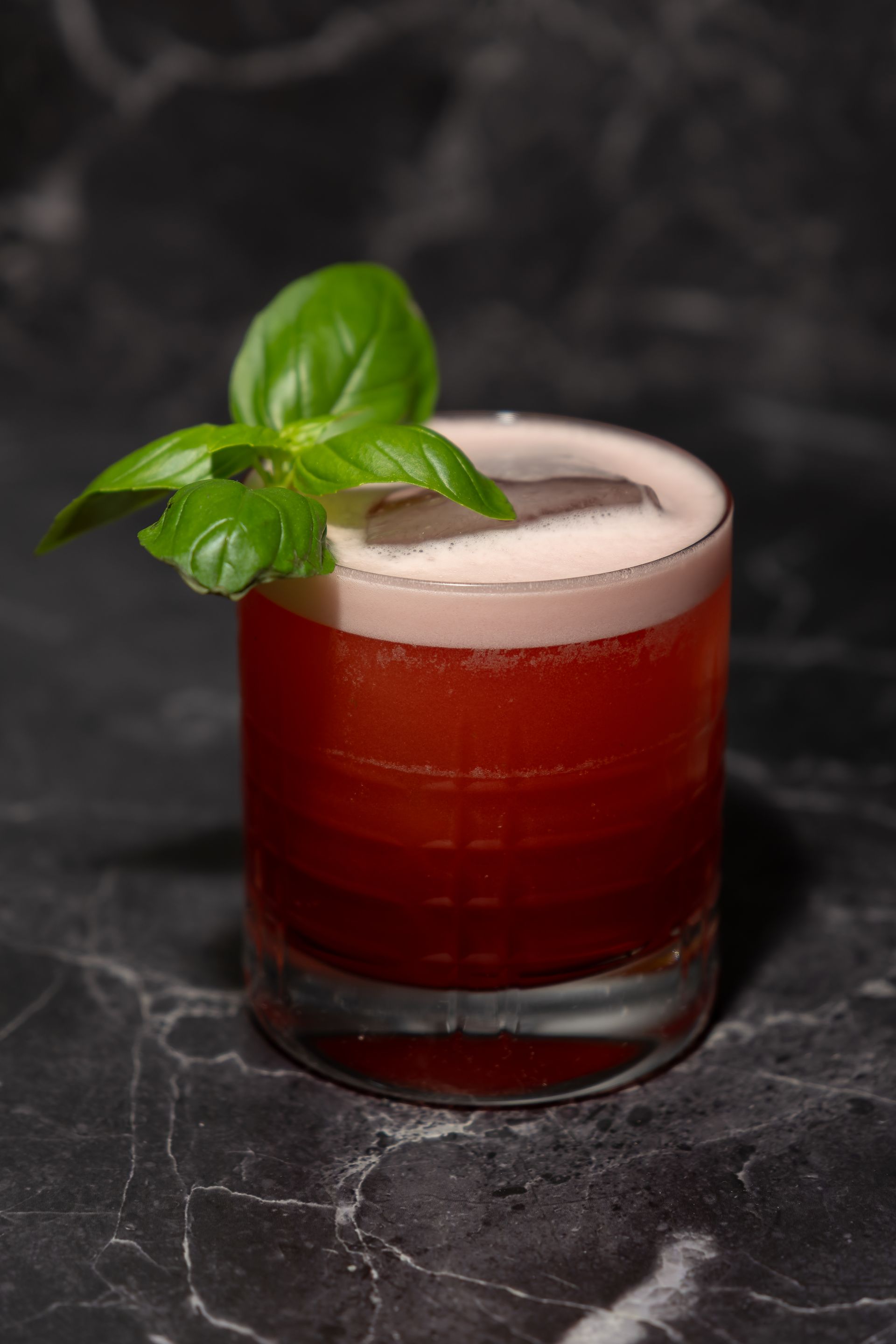 Roter Cocktail in einem niedrigen Glas, garniert mit Basilikum auf einer dunklen Marmoroberfläche