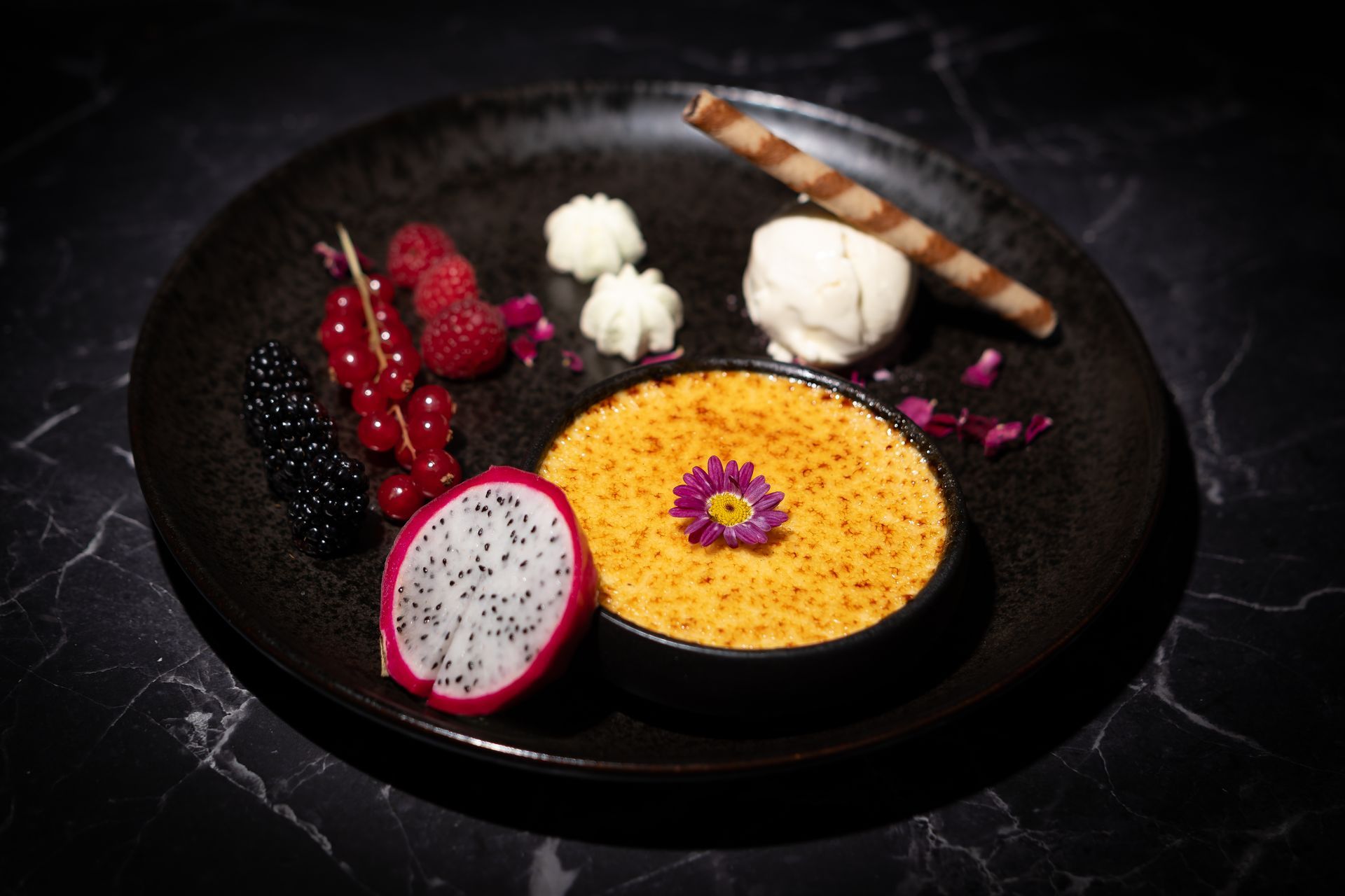 Dessertschale mit gelber Mousse, Drachenfrucht, Beeren, Baiser und Schokoladengarnitur auf einem dunklen Teller