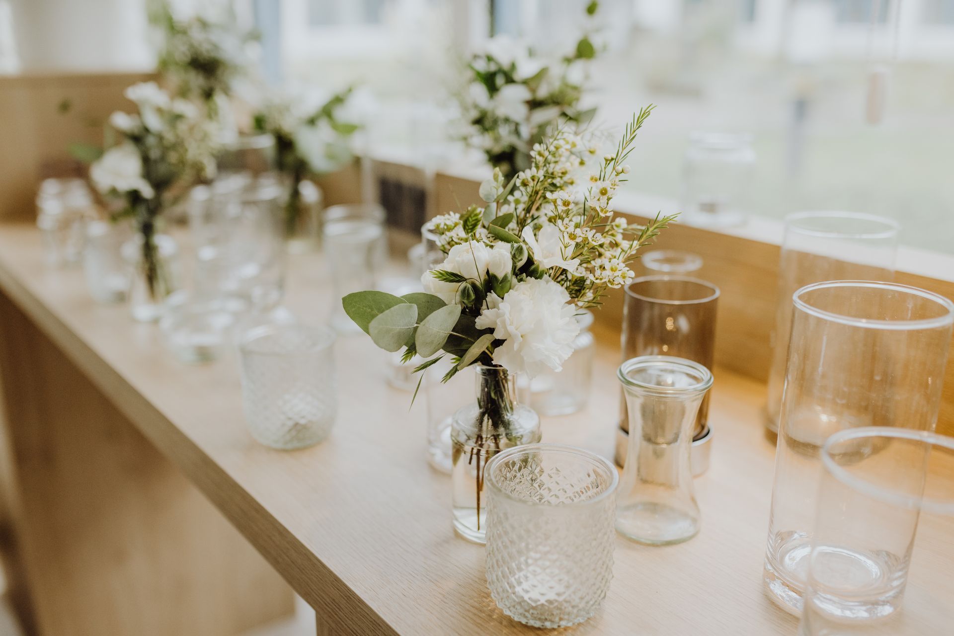 Ein festlich gedeckter Tisch mit Gläsern, Kerzen und kleinen weißen Blumenarrangements in einem hellen Raum