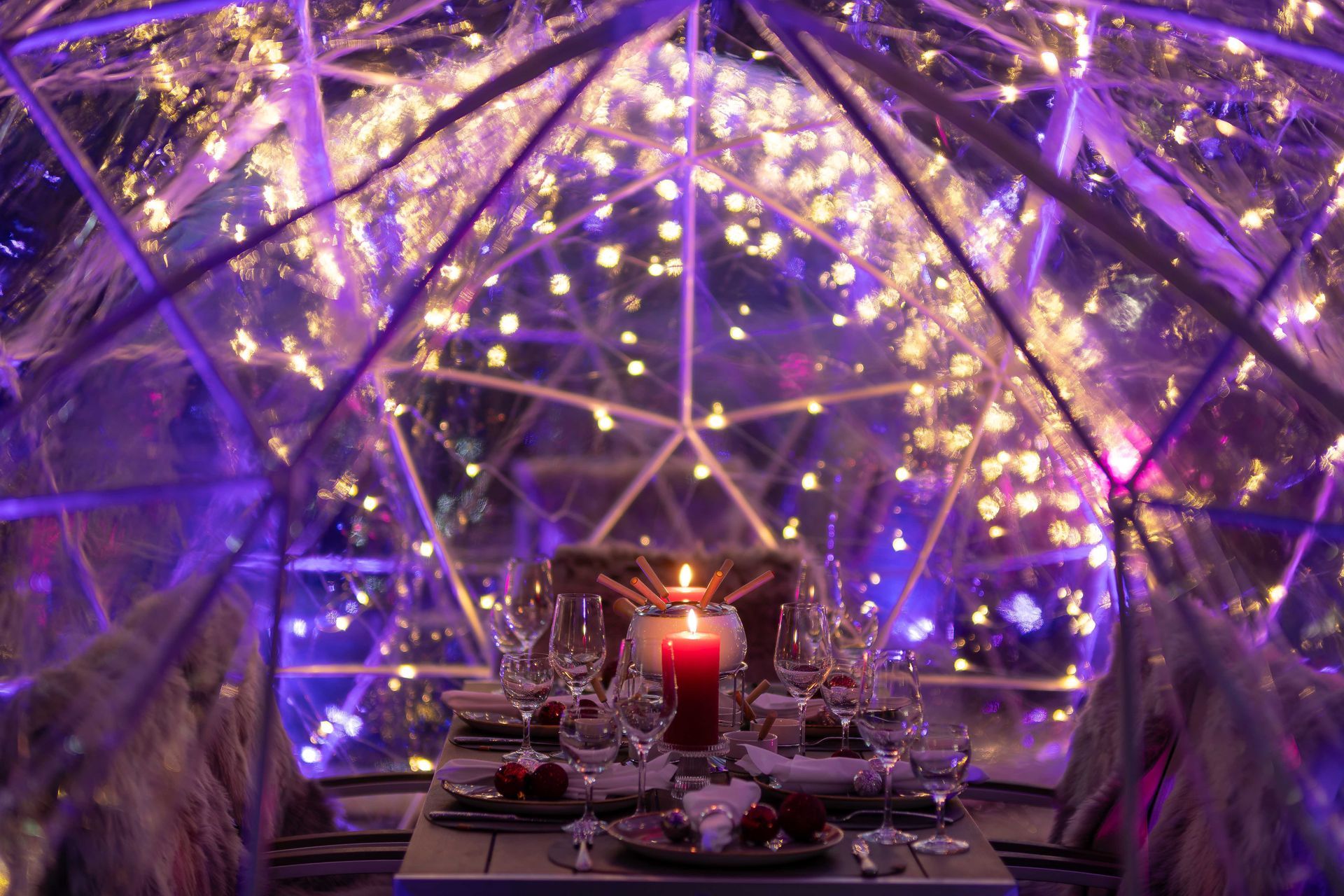 Ein mit Kerzen beleuchteter Esstisch in einer violett erleuchteten Glaskuppel mit funkelnden Lichtern darüber.