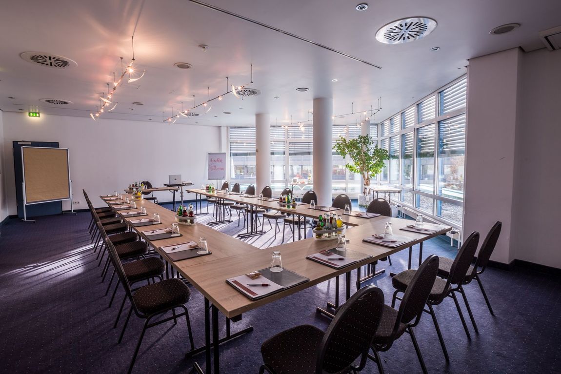 Moderner Konferenzraum mit langen Tischen, schwarzen Stühlen und großen Fenstern mit Blick auf einen hellen Büroraum