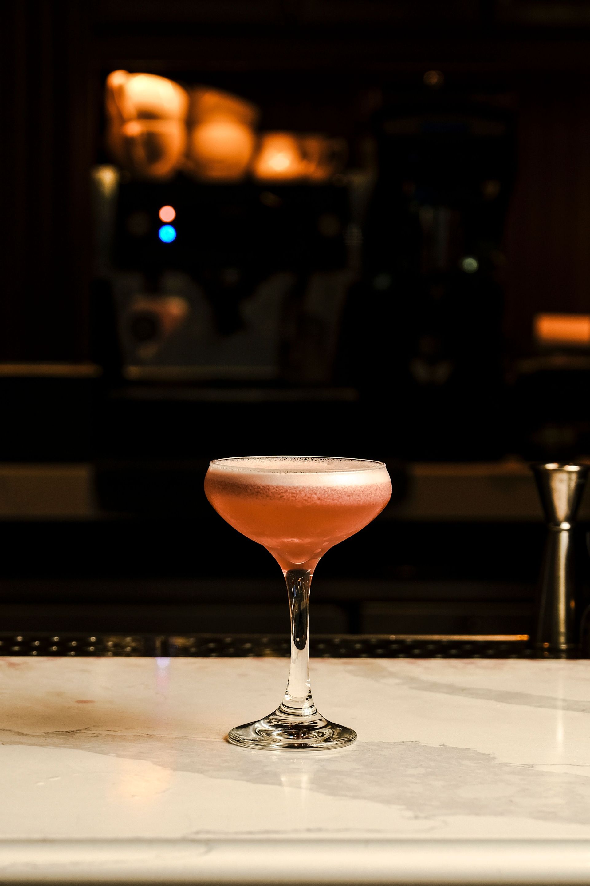 Ein rosafarbener Cocktail in einem Coupé-Glas auf einer Bartheke vor einem verschwommenen, warm beleuchteten Hintergrund.