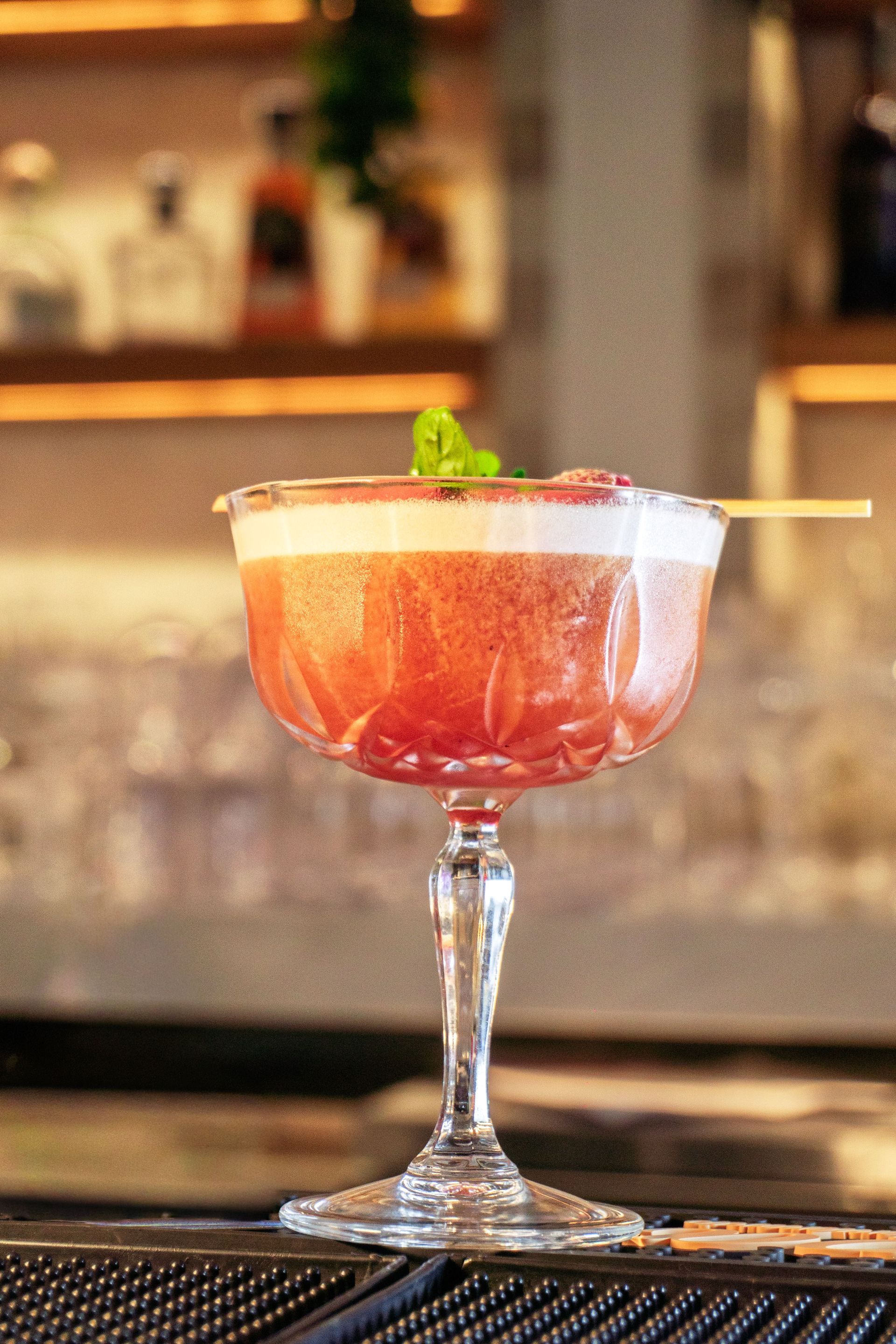 Ein rosafarbener Cocktail in einem Coupé-Glas mit Minzgarnitur auf einer Bartheke