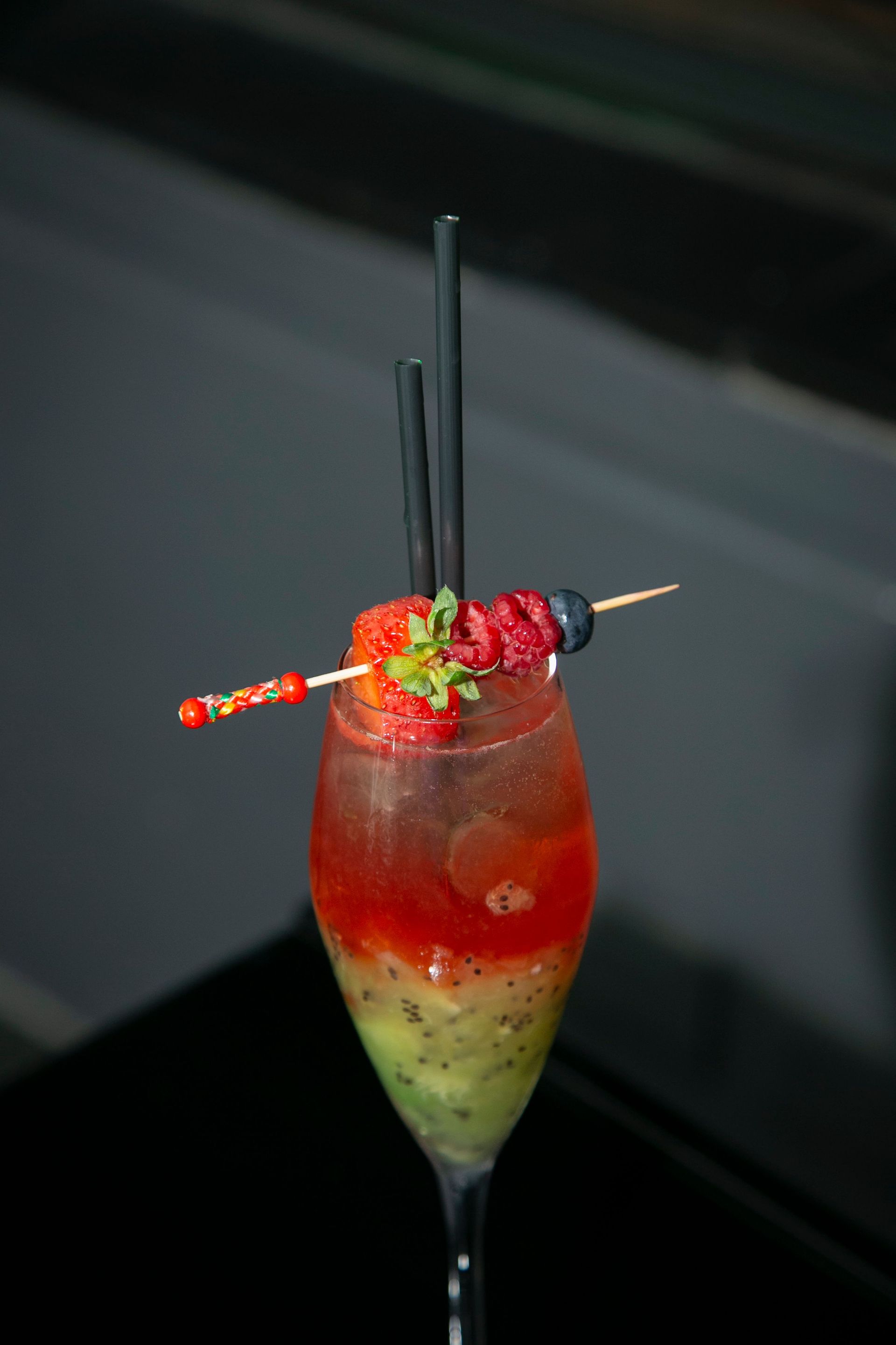 Ein hoher, geschichteter roter und grüner Cocktail, garniert mit Erdbeeren und schwarzen Strohhalmen, serviert in einem Sektglas.