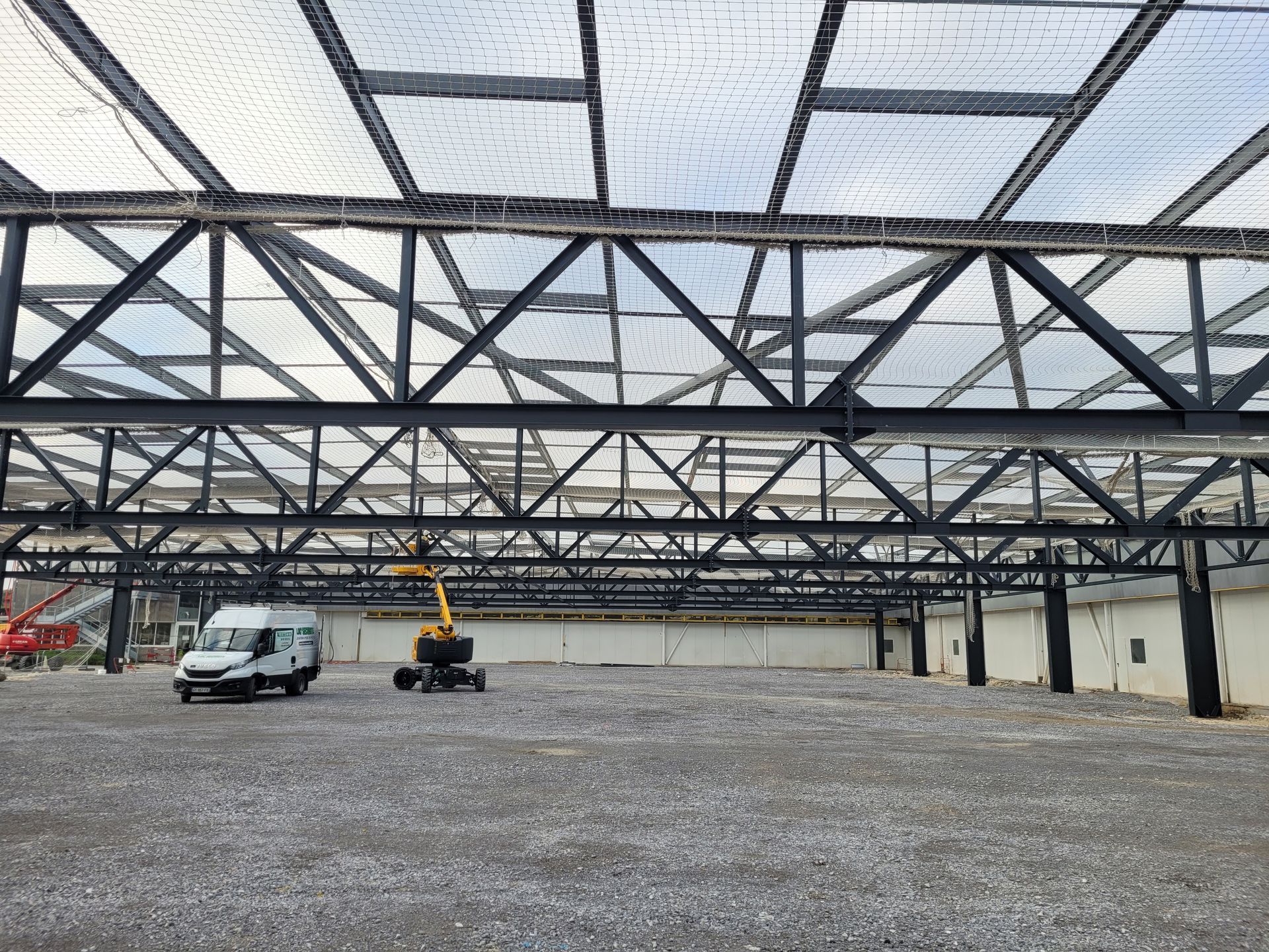 Intérieur d'un grand bâtiment en construction avec poutres d'acier noires apparentes et toiture en filet de sécurité.