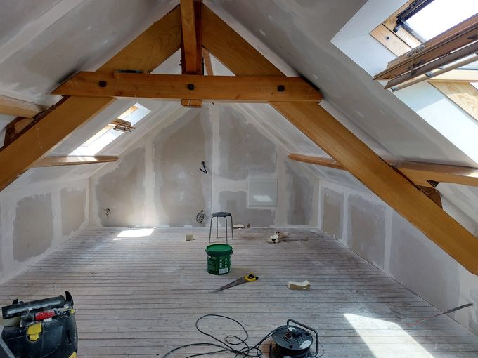Pièce mansardée en cours d'aménagement avec poutres apparentes, murs en plaques de plâtre et plancher en bois brut.