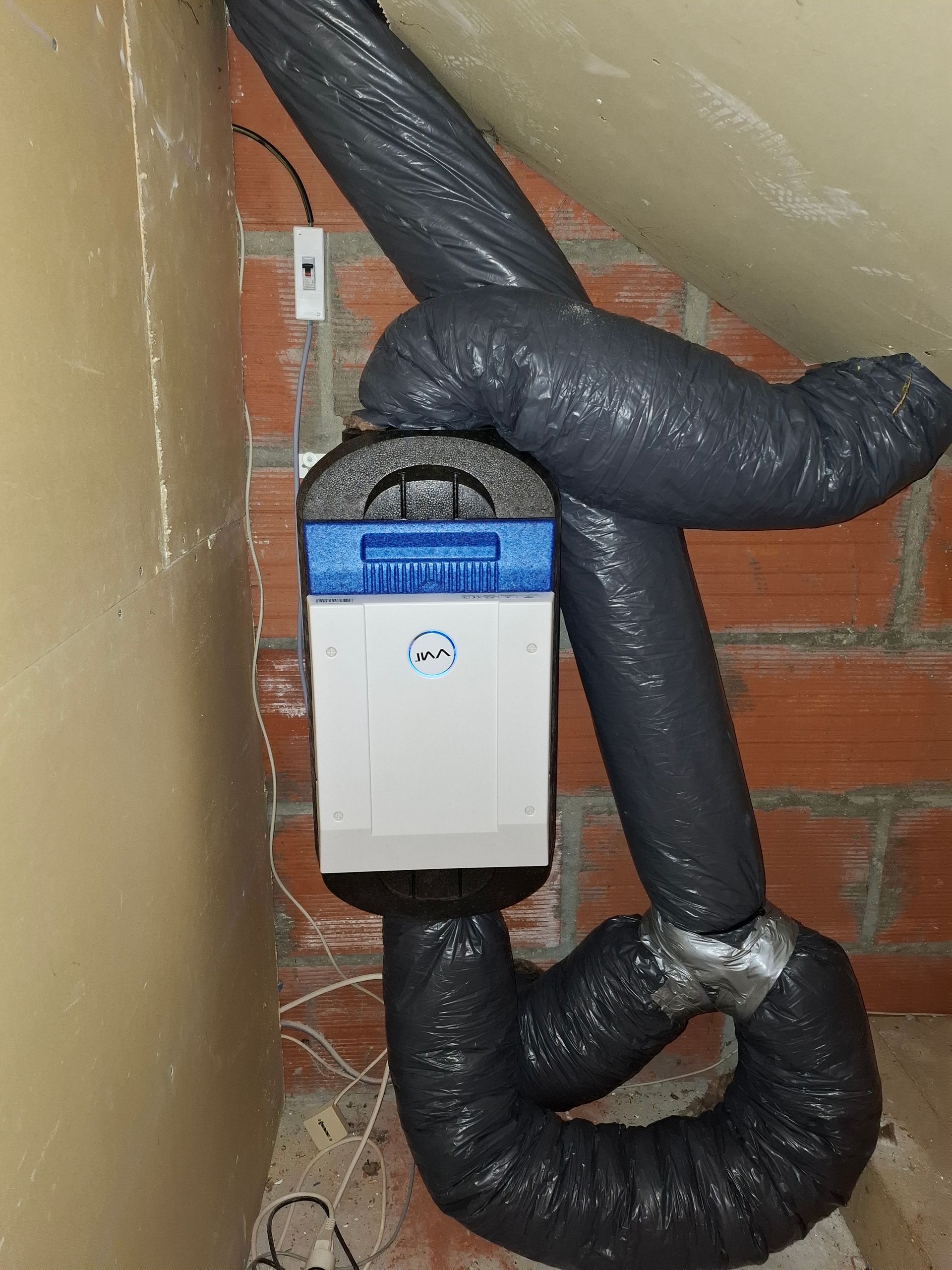 Unité de ventilation blanche et bleue avec conduits gris flexibles dans un espace aux murs de briques.