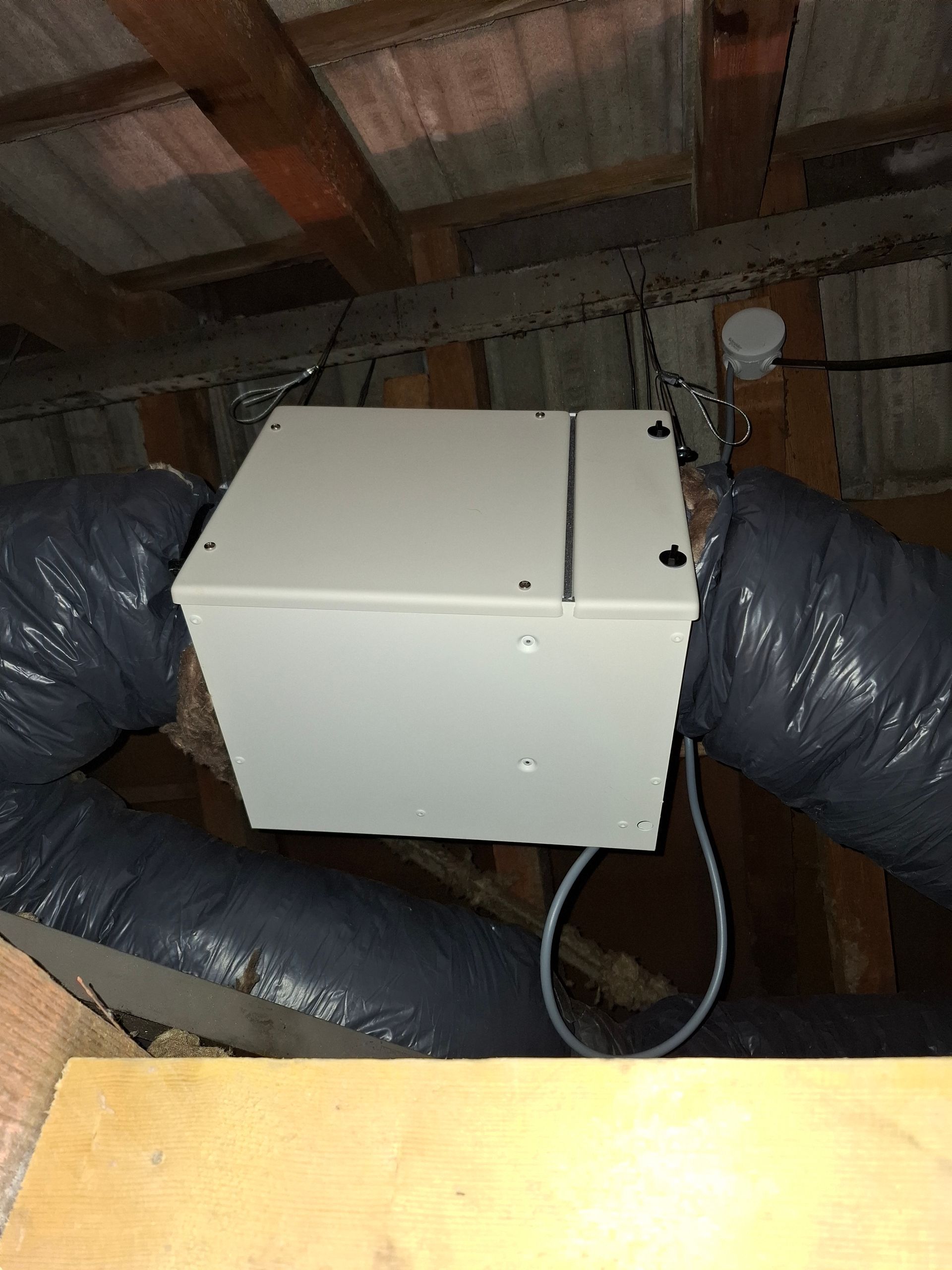 Une unité de climatisation rectangulaire grise suspendue dans un grenier, reliée par des conduits flexibles.