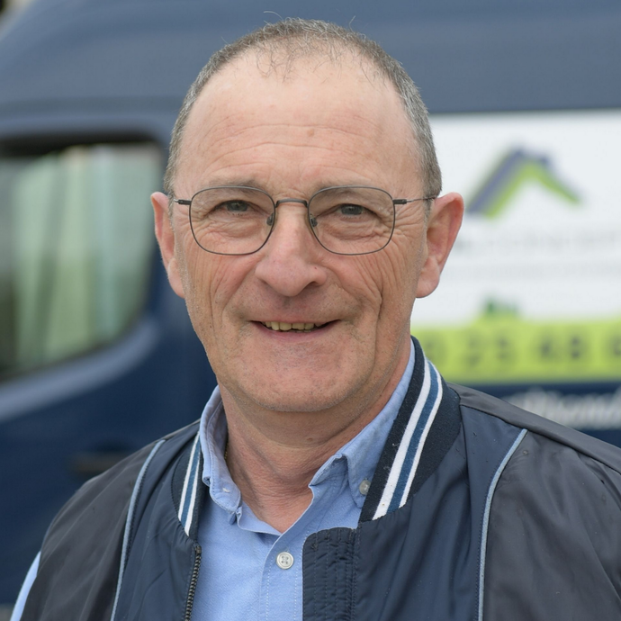 Un homme souriant, portant des lunettes et une veste bleue, se tient devant un véhicule bleu.