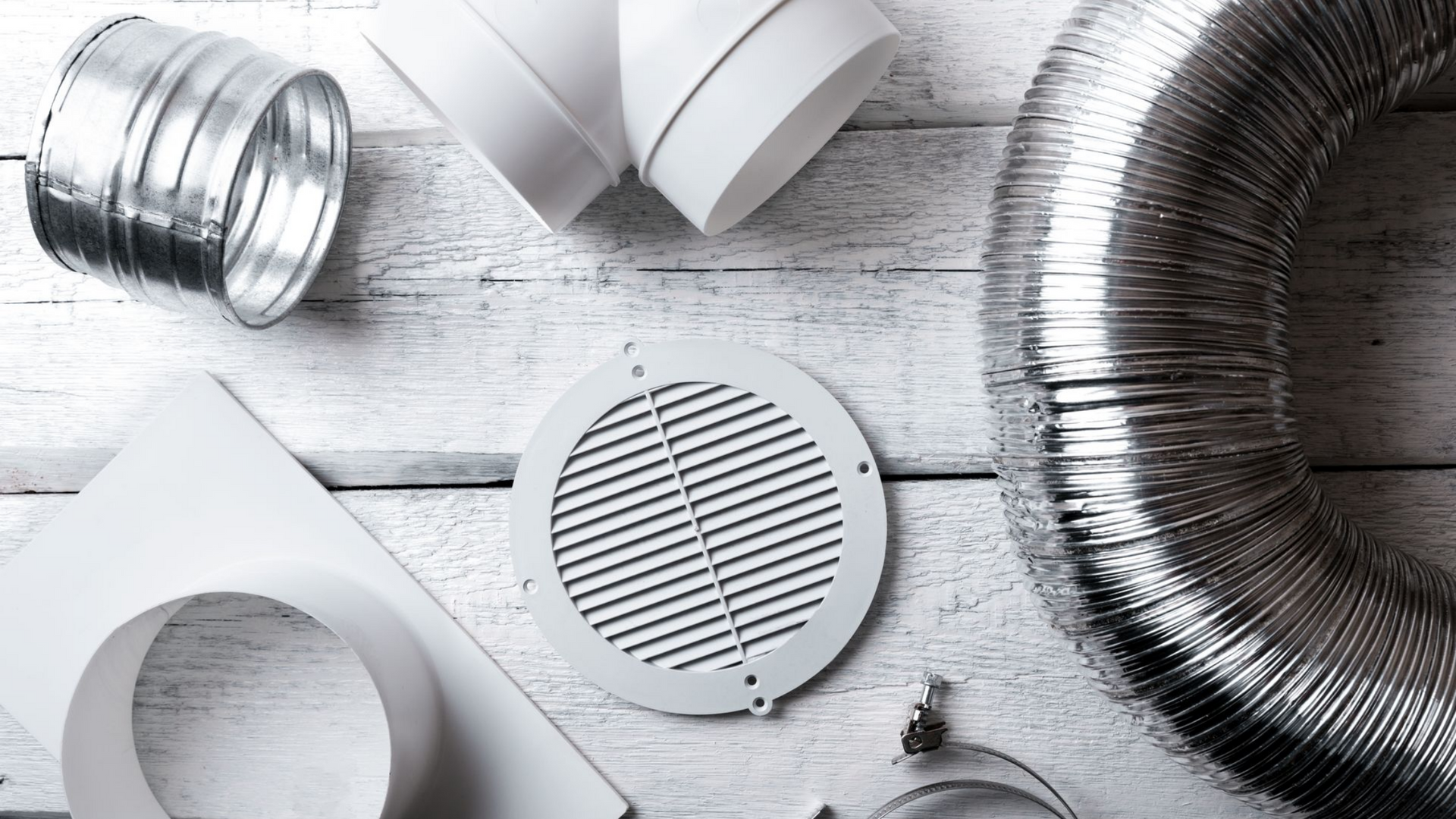 Éléments du système de ventilation : conduits métalliques et blancs, grille d’aération et tuyauterie flexible sur une surface en bois blanc.