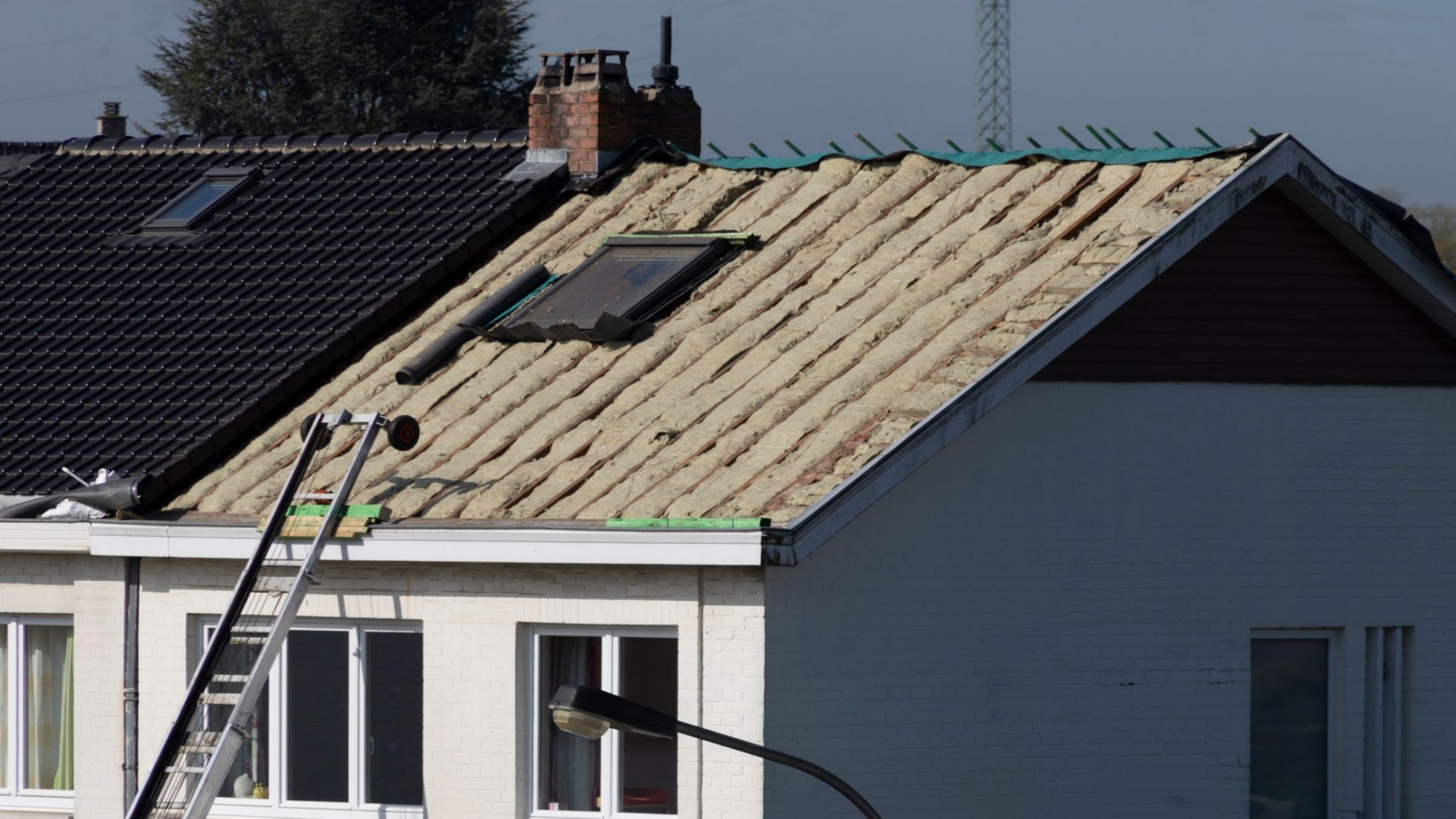 Maison à la toiture partiellement refaite ; sous-toiture et tuiles apparentes. Une échelle est appuyée contre la maison.