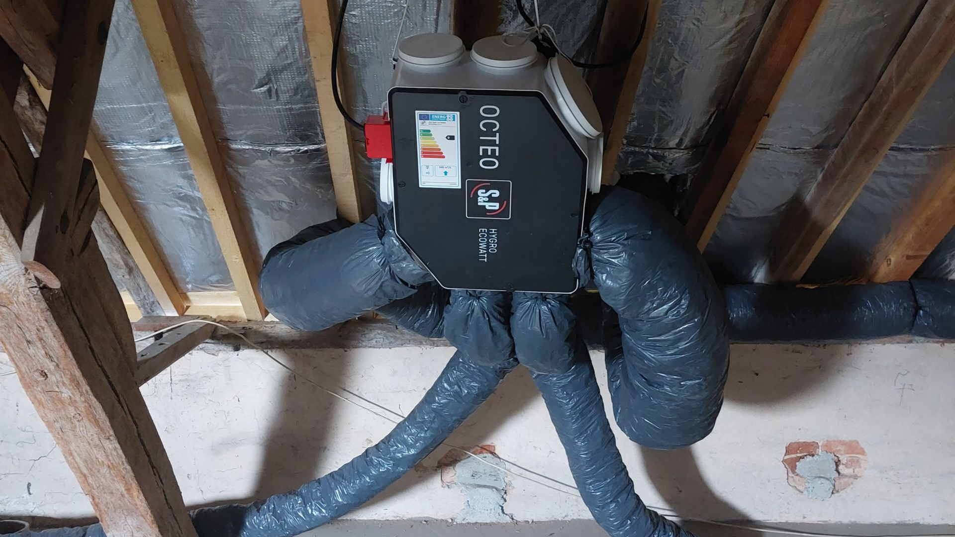 Une unité de ventilation installée dans un grenier avec des conduits isolés et des poutres en bois.