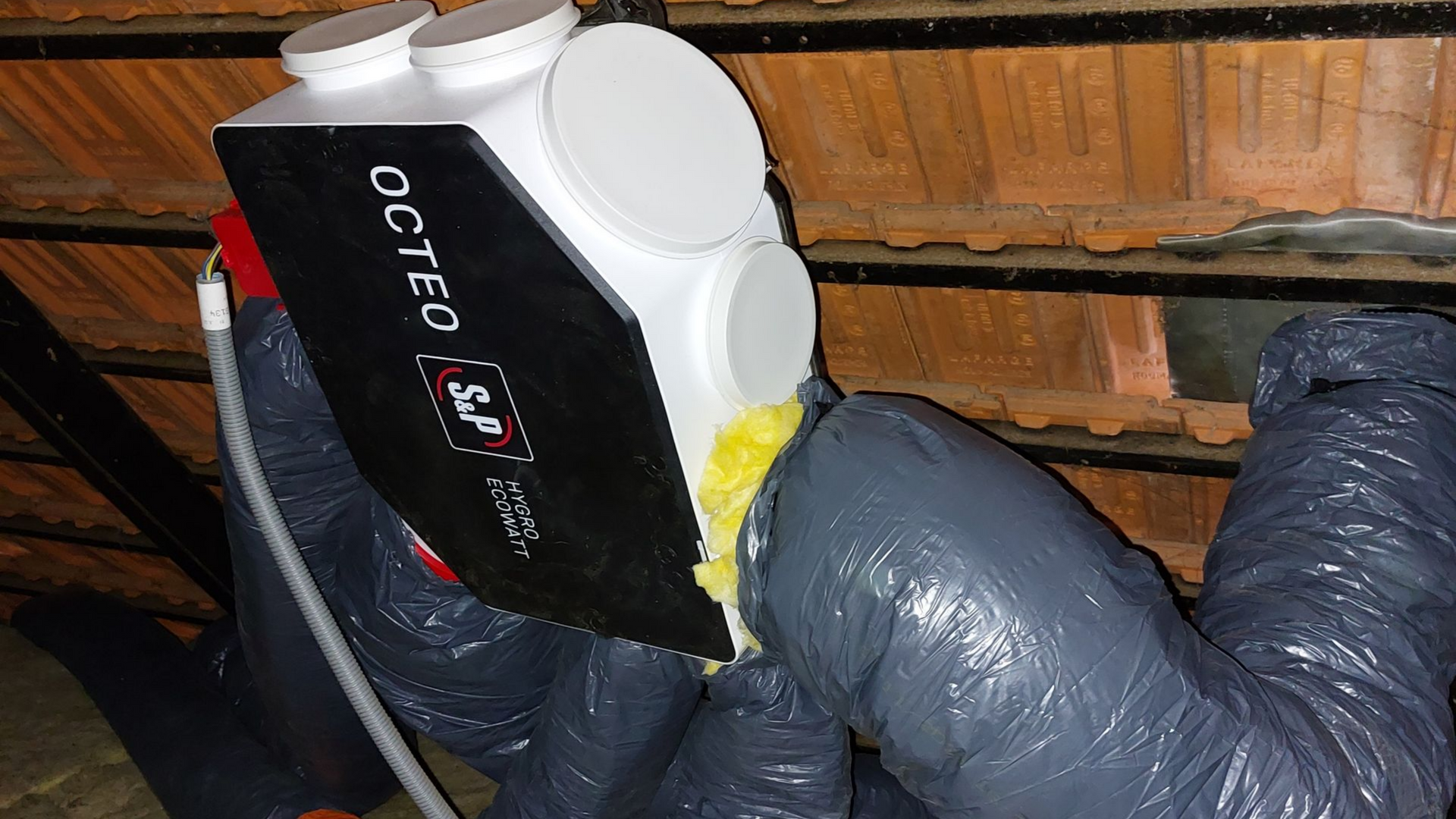 Un système de ventilation de combles, installé dans des combles revêtus de briques. Des conduits isolés gris sont raccordés à l'unité.