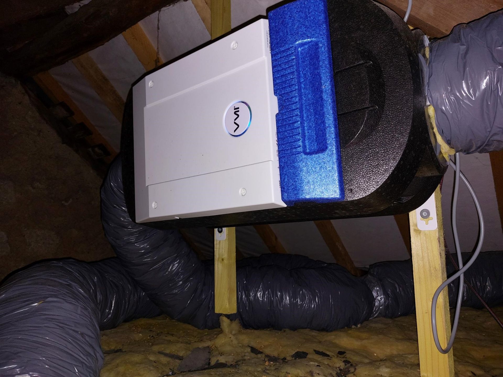 Unité de déshumidification dans un grenier, avec conduits flexibles et supports en bois, à côté de l'isolant.
