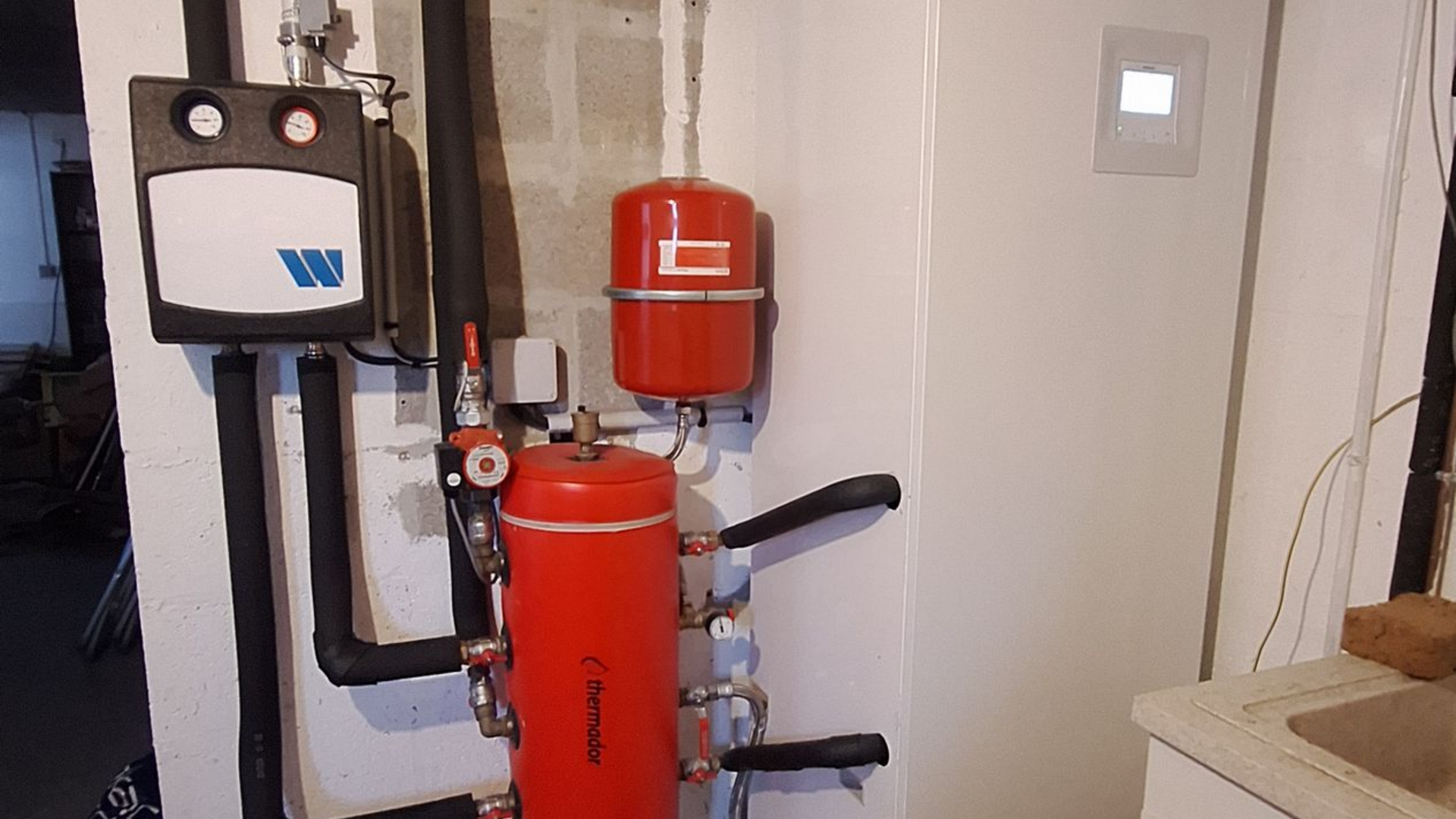 Système de chauffage avec réservoirs rouges, tuyaux noirs, panneau de commande blanc et mur blanc.