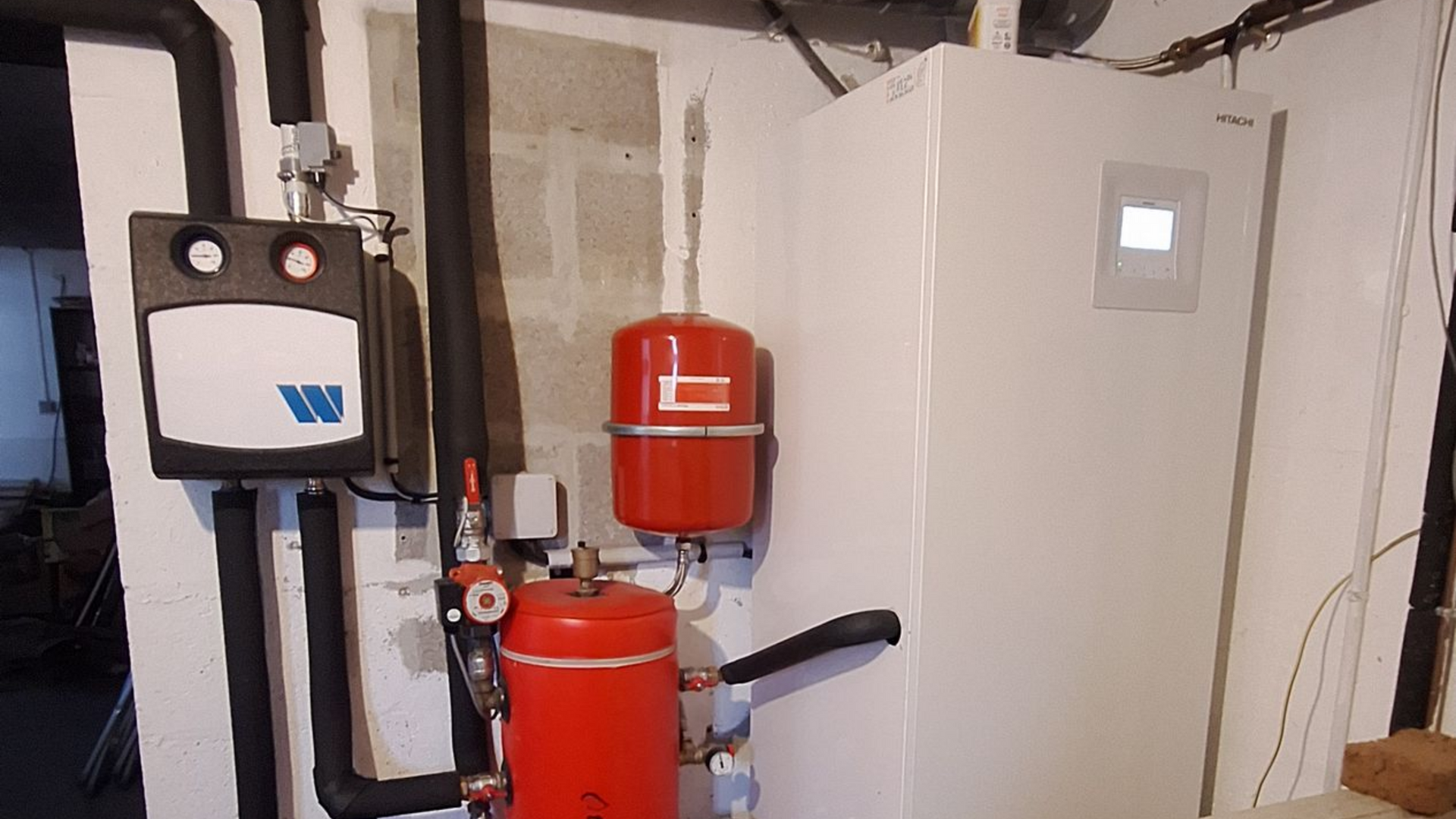 Pompe à chaleur blanche et vases d'expansion rouges dans une buanderie, avec tuyaux isolés noirs et panneau de commande.