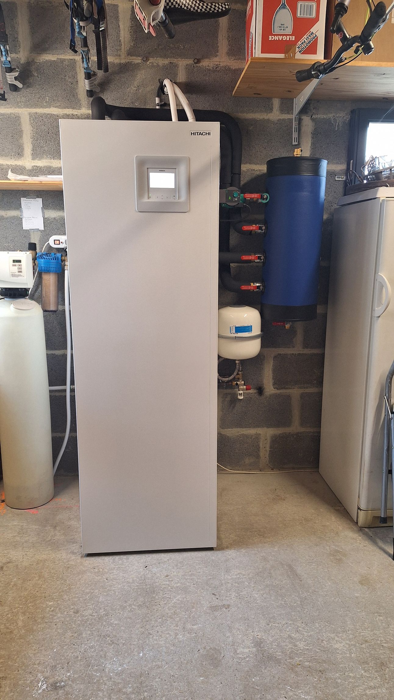 Grande unité de chauffage grise contre un mur en parpaings, avec un réservoir d'eau bleu et d'autres appareils de plomberie.