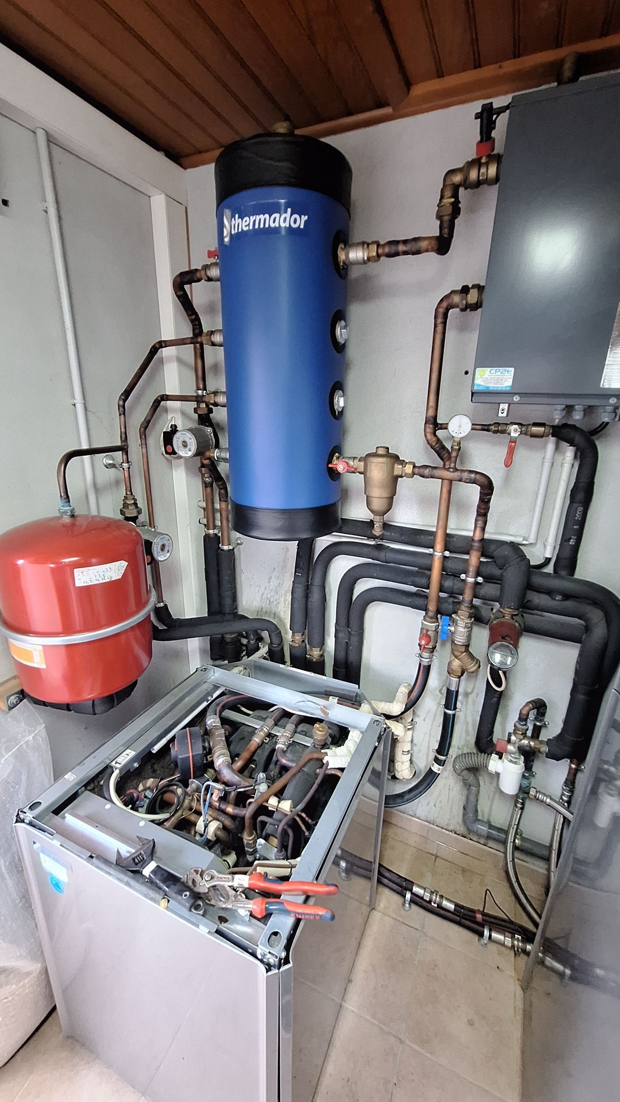 Chauffe-eau bleu, vase d'expansion rouge et tuyaux complexes dans une buanderie.