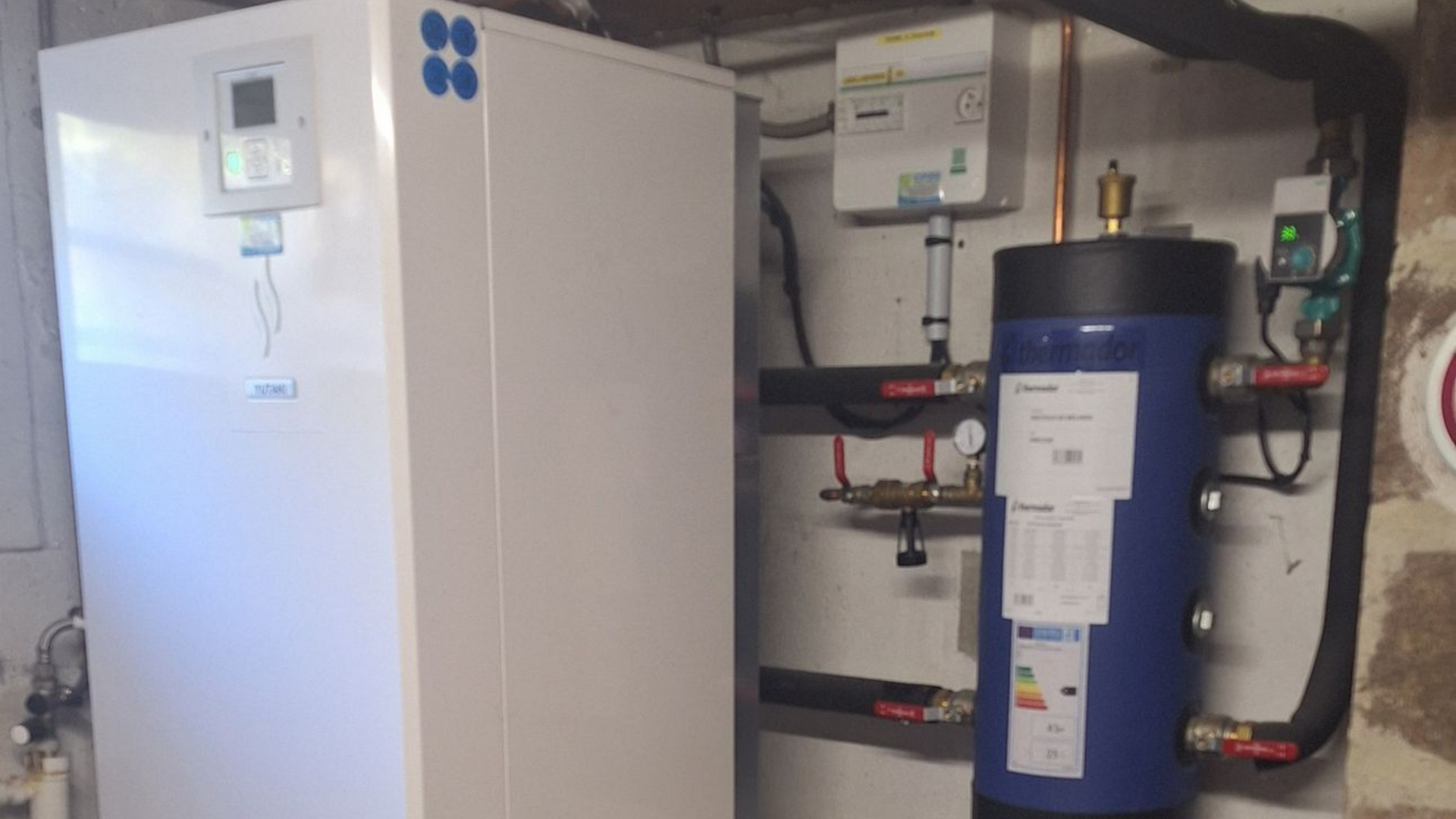 Pompe à chaleur blanche à côté d'un chauffe-eau bleu dans une buanderie avec tuyaux et composants électriques.