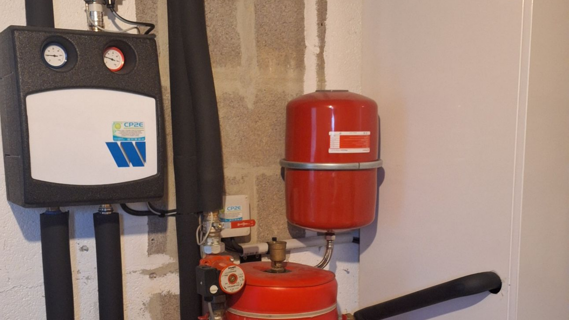 Système de chauffage mural avec boîtier de commande noir et vase d'expansion rouge sur mur en béton.