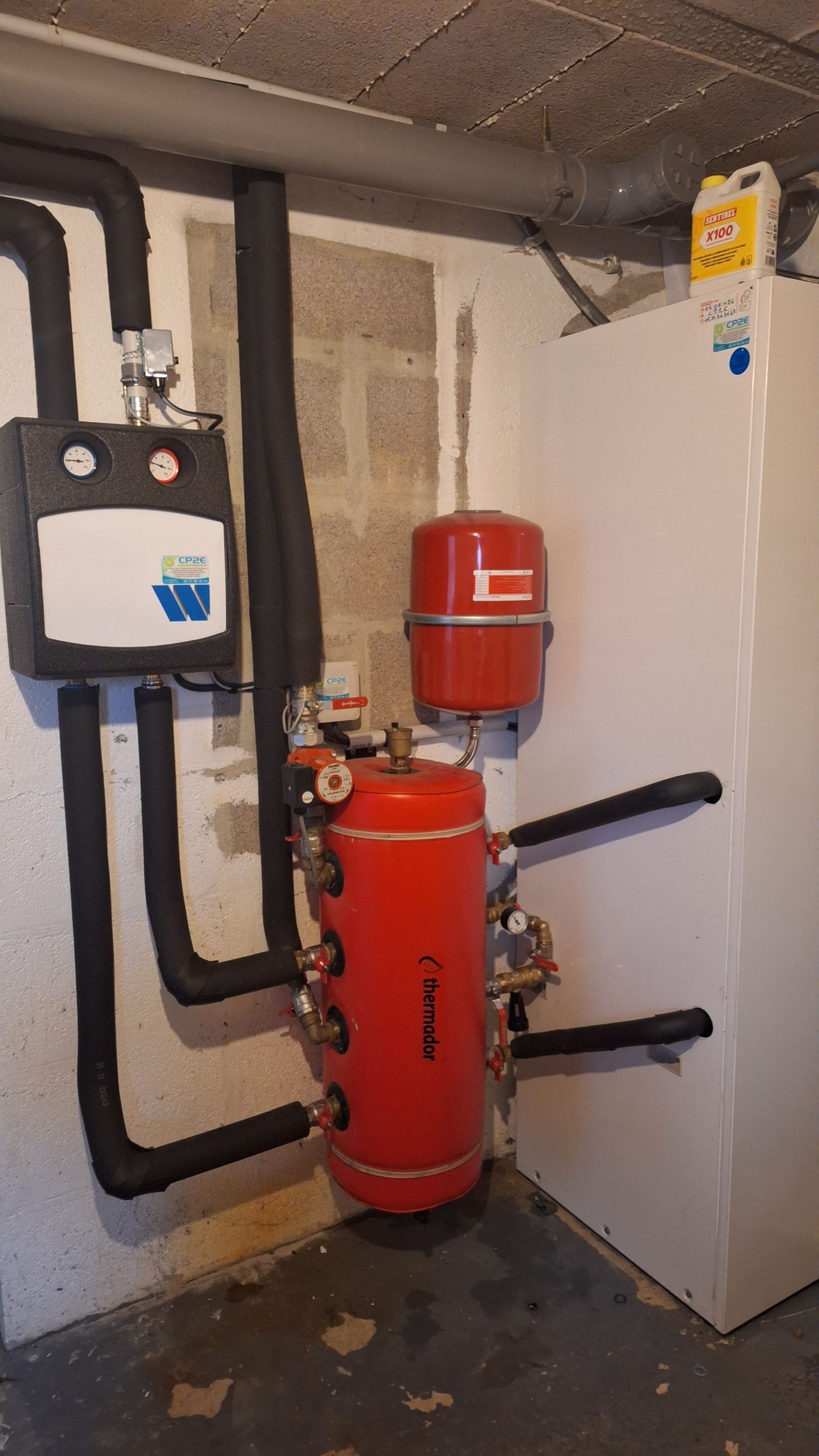 Système de chauffage rouge, avec tuyaux noirs et réservoir d'eau blanc dans un coin du sous-sol.