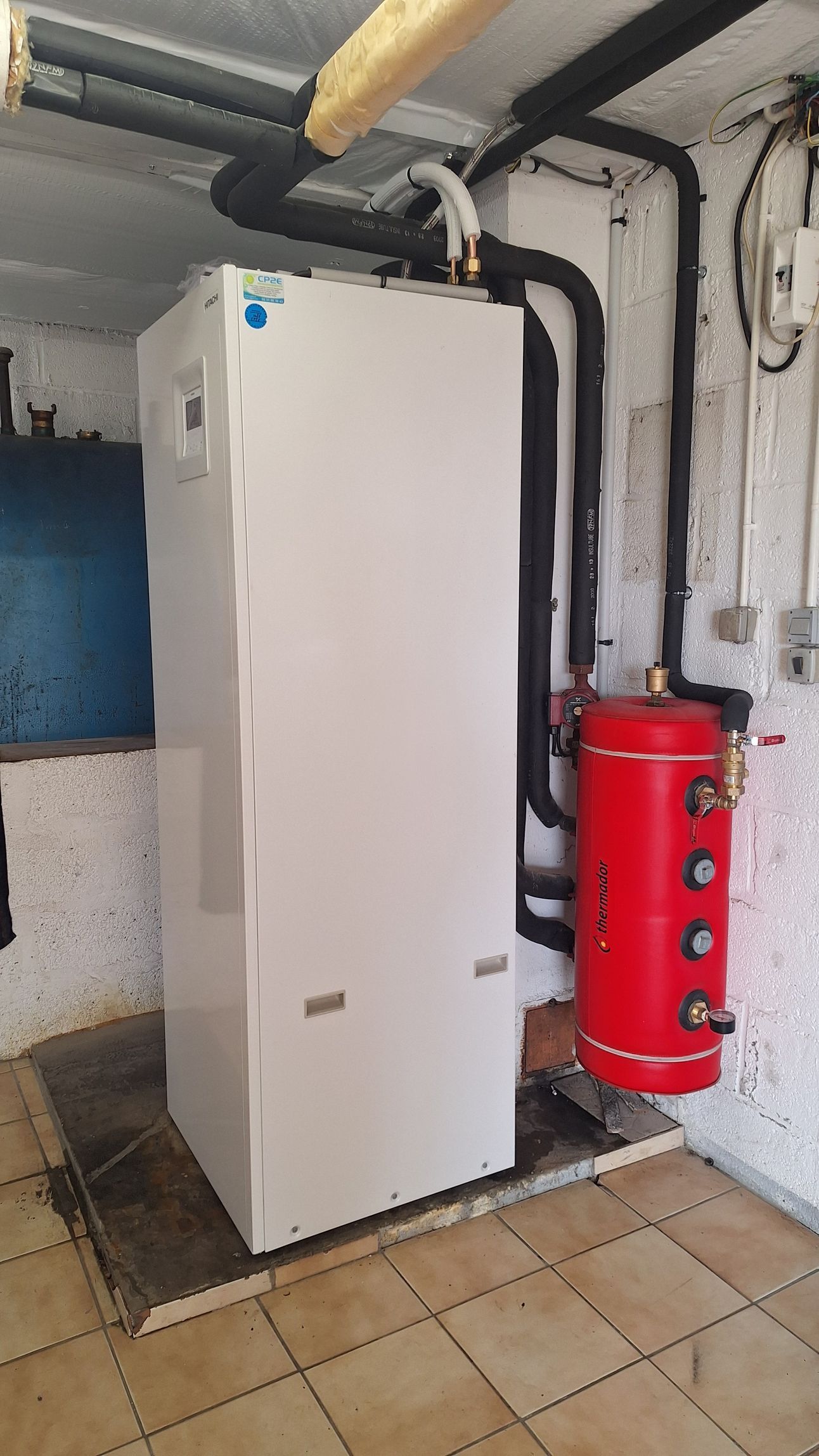 Pompe à chaleur blanche et chauffe-eau rouge dans une pièce. Tuyaux noirs, sol beige, mur en briques.