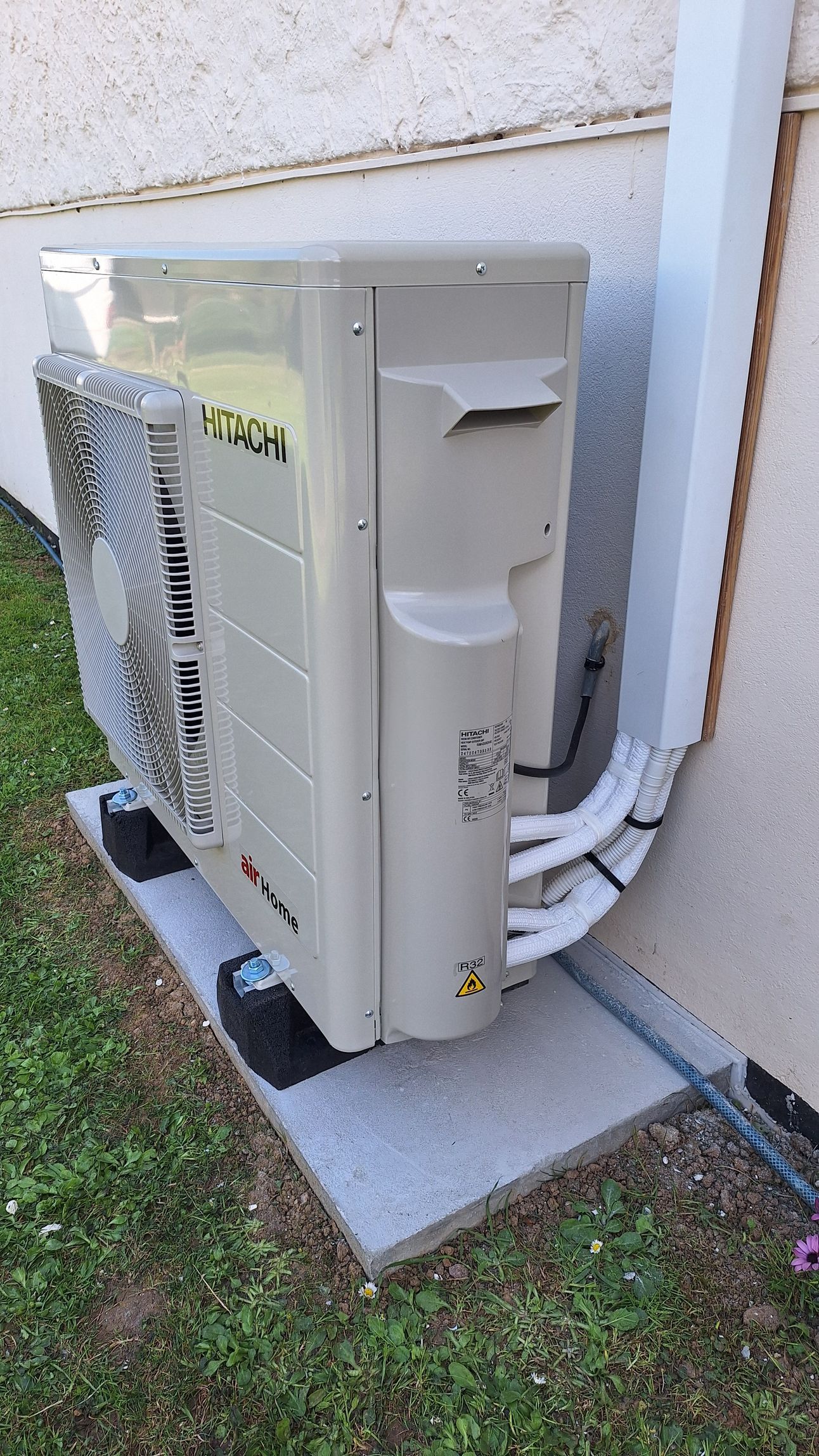 Unité de climatisation extérieure, beige et argent, sur une dalle de béton à côté d'un mur blanc et d'herbe verte.