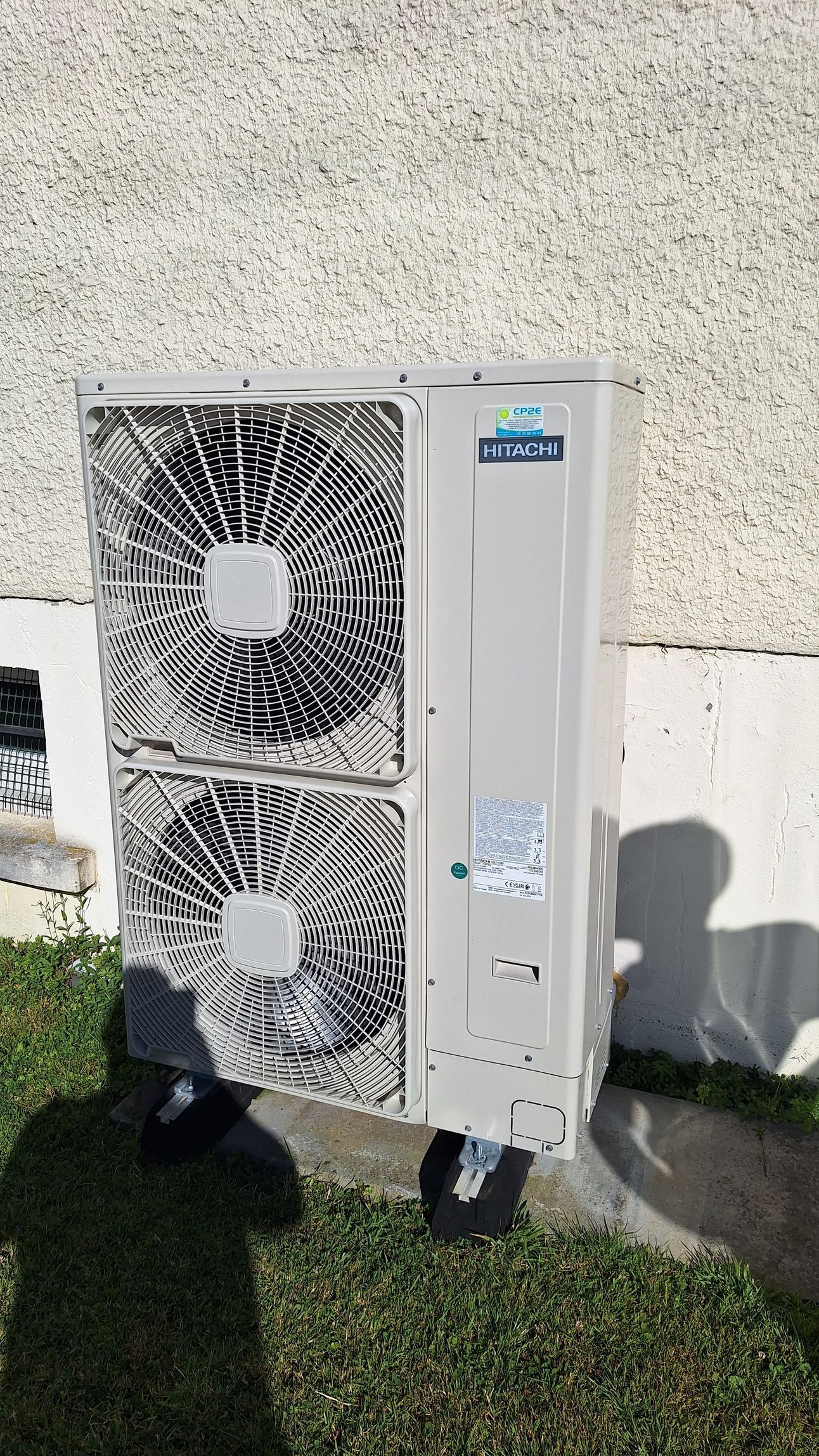 Climatiseur fixé au mur, près de la pelouse. Gris et blanc, avec ventilateurs visibles.