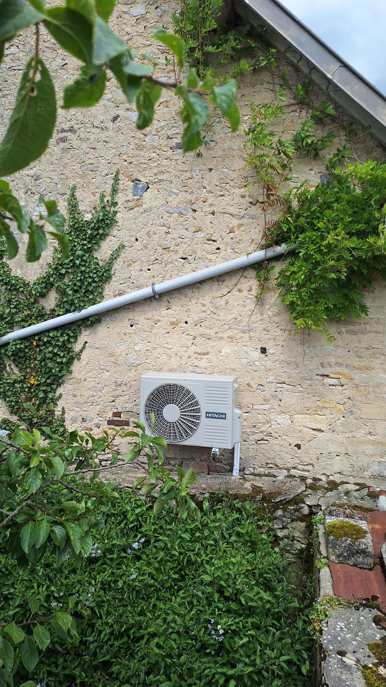 Unité de climatisation sur mur texturé, entourée de feuillage vert et d'un tuyau métallique.