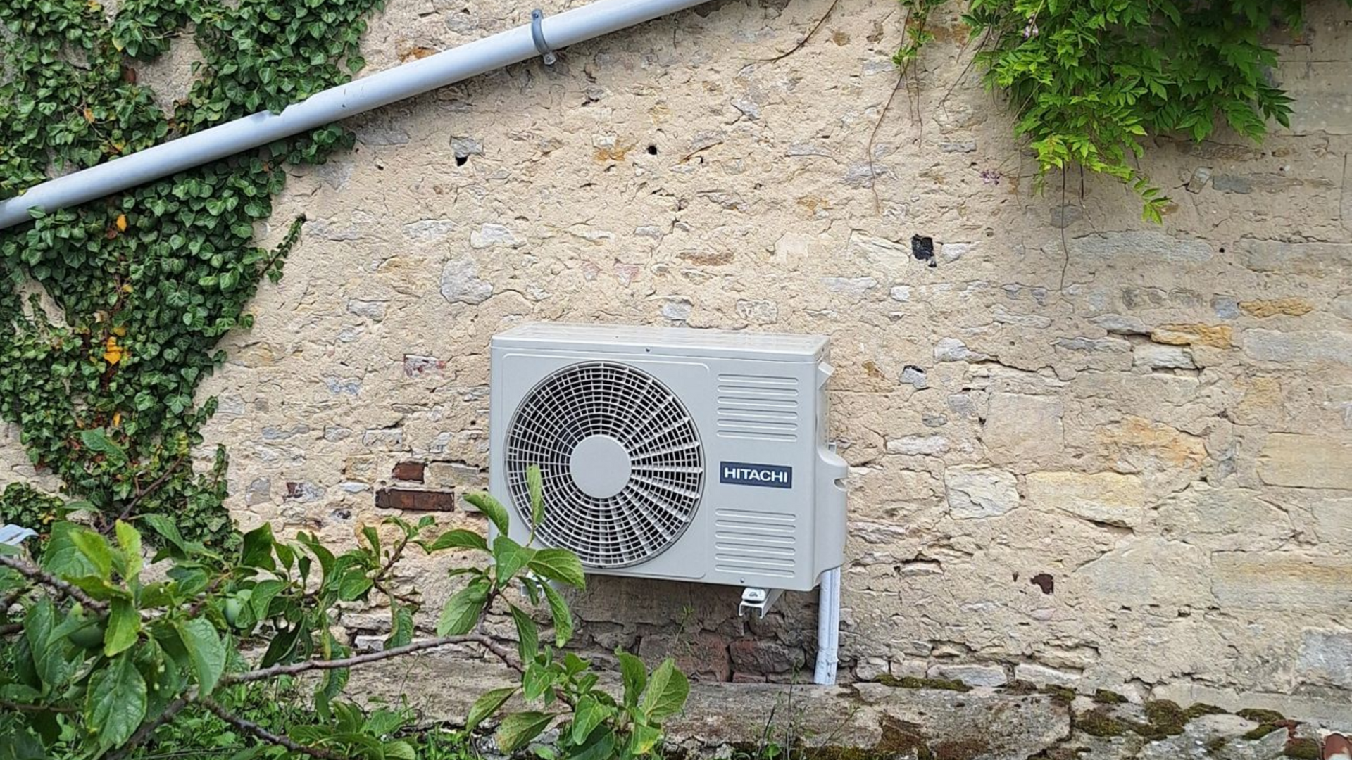 Une unité de climatisation extérieure sur un mur en pierre brute, avec des vignes et un tuyau gris au-dessus.