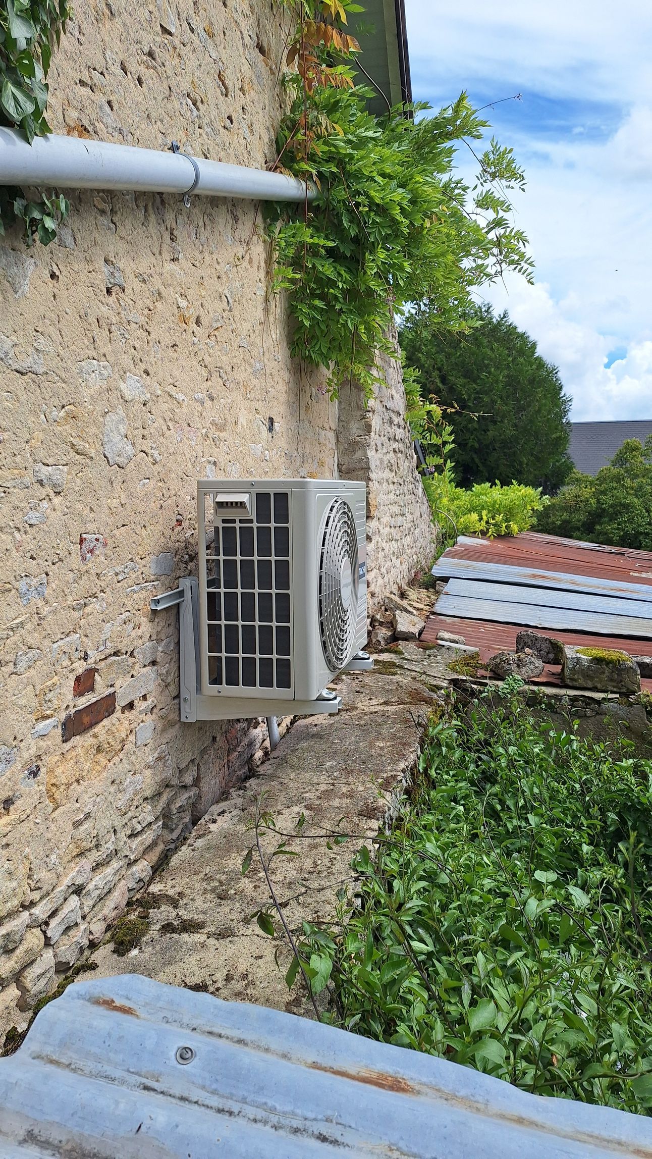 Unité de climatisation sur un mur de pierre à côté de vignes et d'un toit en métal.