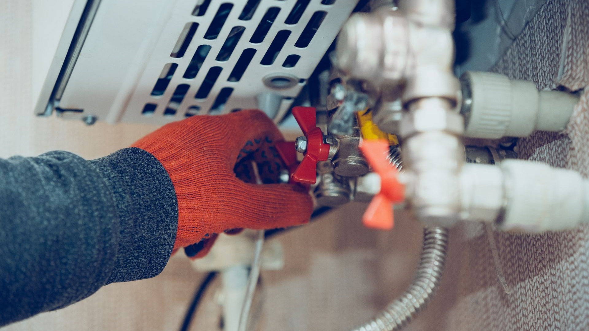 Une main gantée tourne une vanne rouge sur une chaudière avec des tuyaux et des évents.