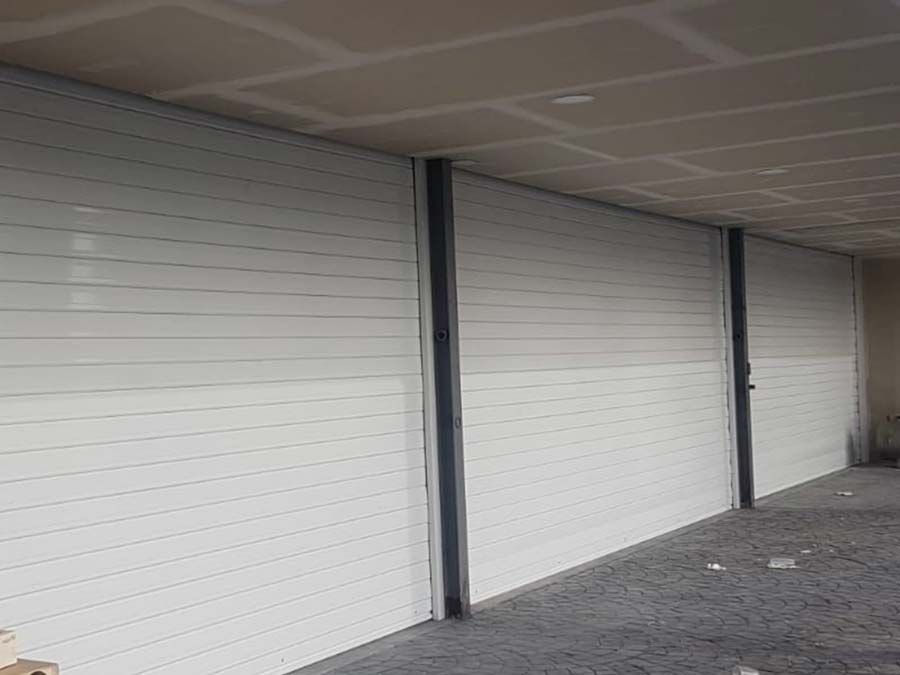 Puertas enrollables blancas a lo largo de una pasarela exterior cubierta con columnas de soporte oscuras.
