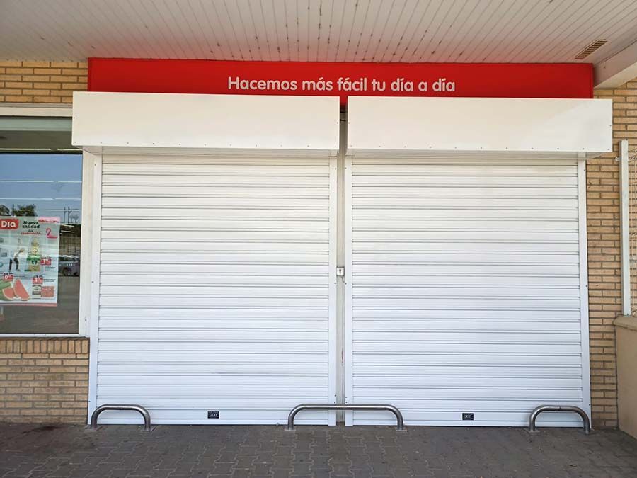 Persianas blancas cerradas en el escaparate de una tienda, bajo un letrero rojo frente a un edificio.