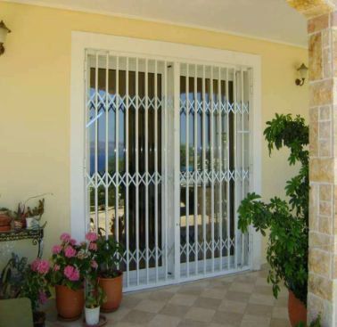 Puerta corrediza blanca con barrotes en un patio soleado con macetas de flores y plantas.