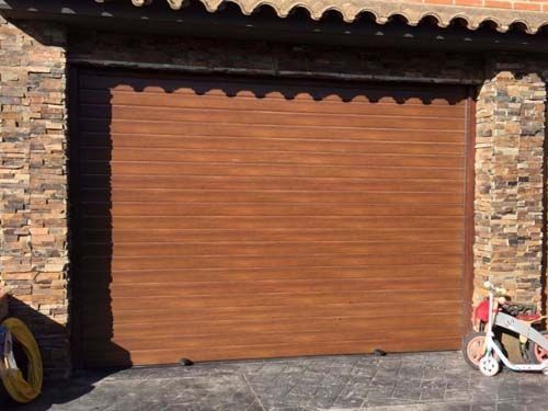 Puerta de garaje marrón cerrada con paredes de piedra y una bicicleta infantil blanca y roja en el borde derecho.