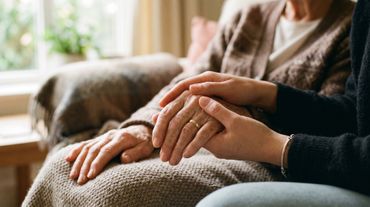 Deux personnes se tenant la main sur un canapé, l'une d'elles posant sa main sur ses genoux recouverts d'une couverture.