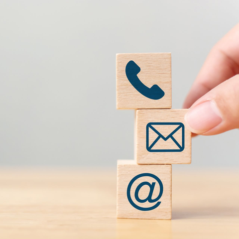 Des blocs de bois empilés, ornés d'icônes de téléphone, d'e-mail et de symbole @, sont posés sur une table. Une main dépose un bloc.