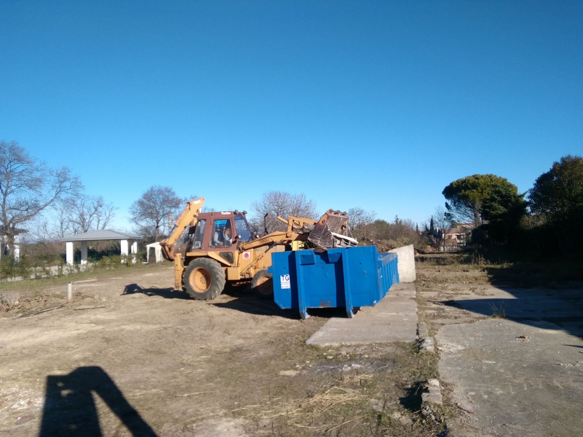 Une pelleteuse jaune décharge des conteneurs bleus sur un terrain vague sous un ciel bleu azur.